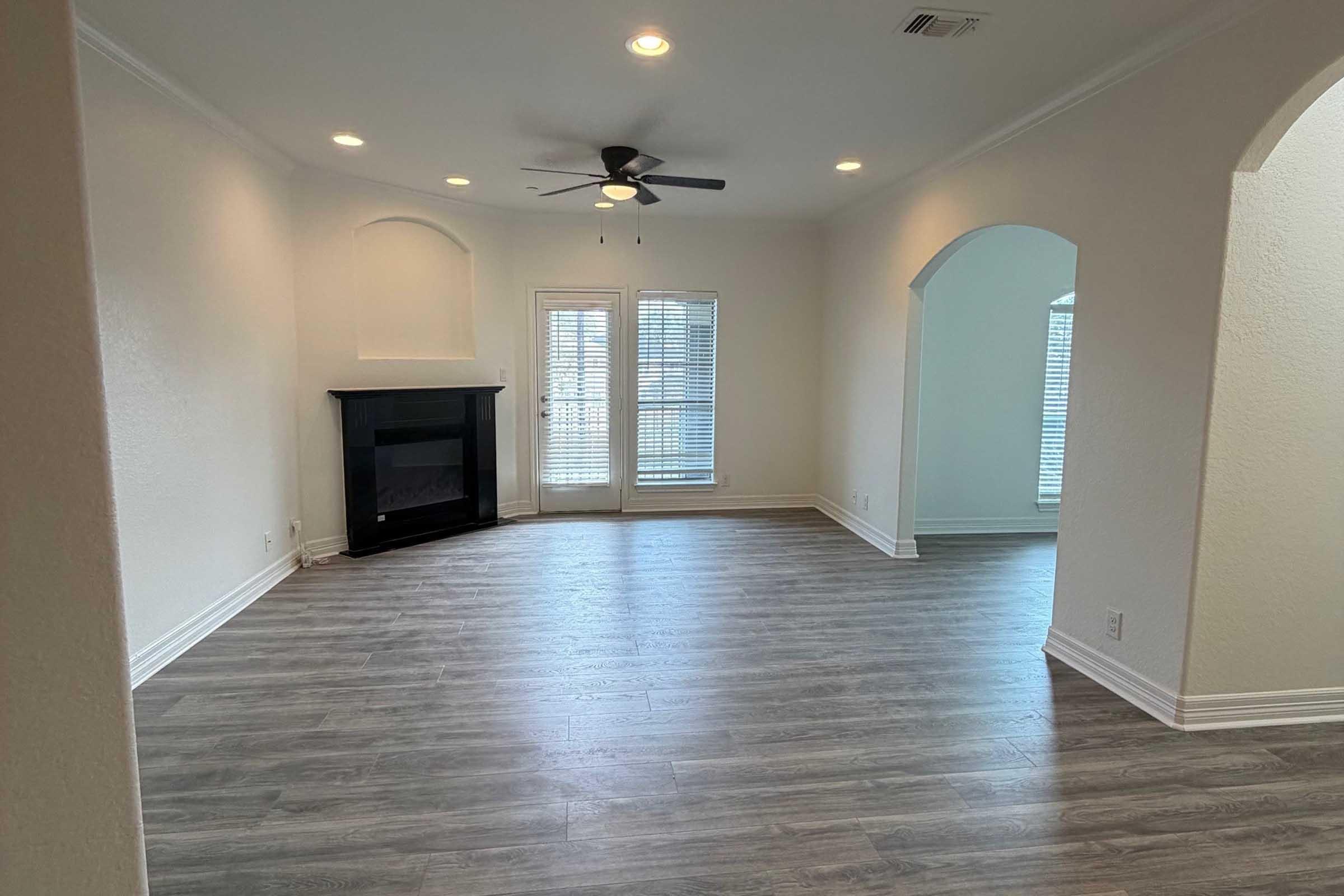 A spacious, empty living room featuring light-colored walls, a ceiling fan, and a cosmetic fireplace. Large windows with blinds allow natural light to fill the space, which has a modern wooden floor. Arched doorways lead to other areas, creating an open and airy atmosphere.