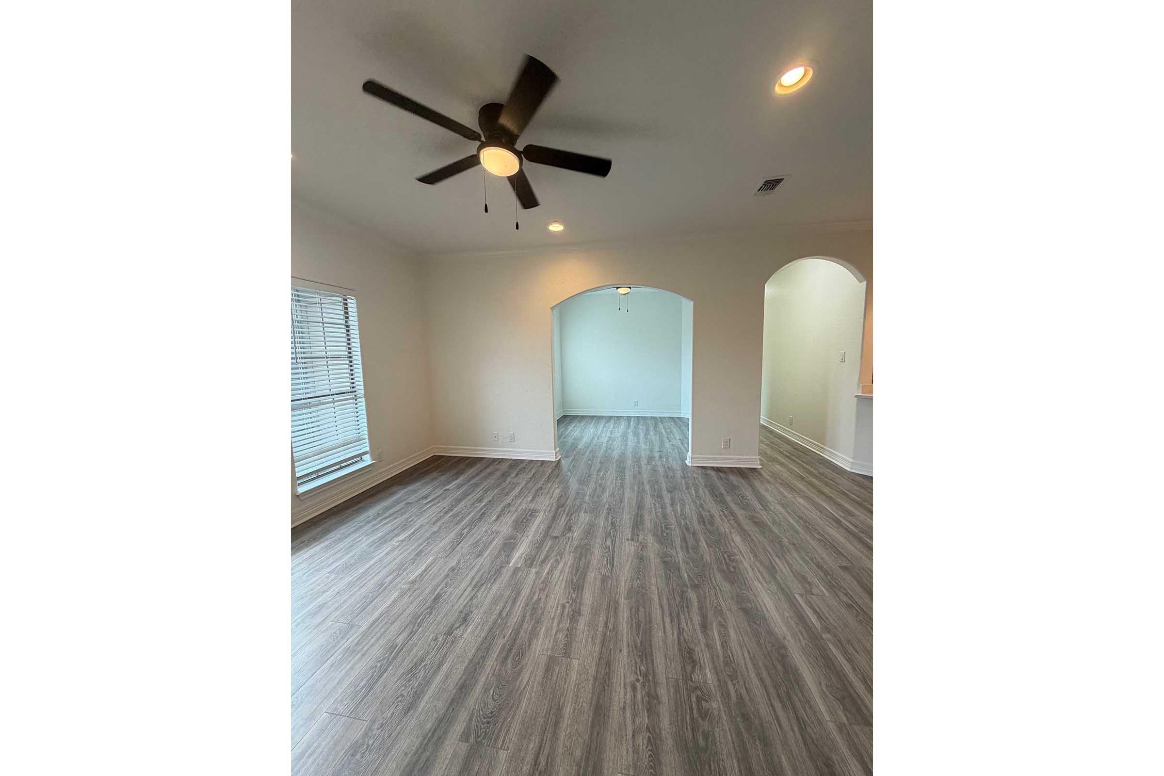 A spacious living area with light gray wood flooring, featuring a ceiling fan. Natural light comes in through a large window with blinds. An archway leads to another room, and there are neutral-colored walls. The overall ambiance is bright and inviting.