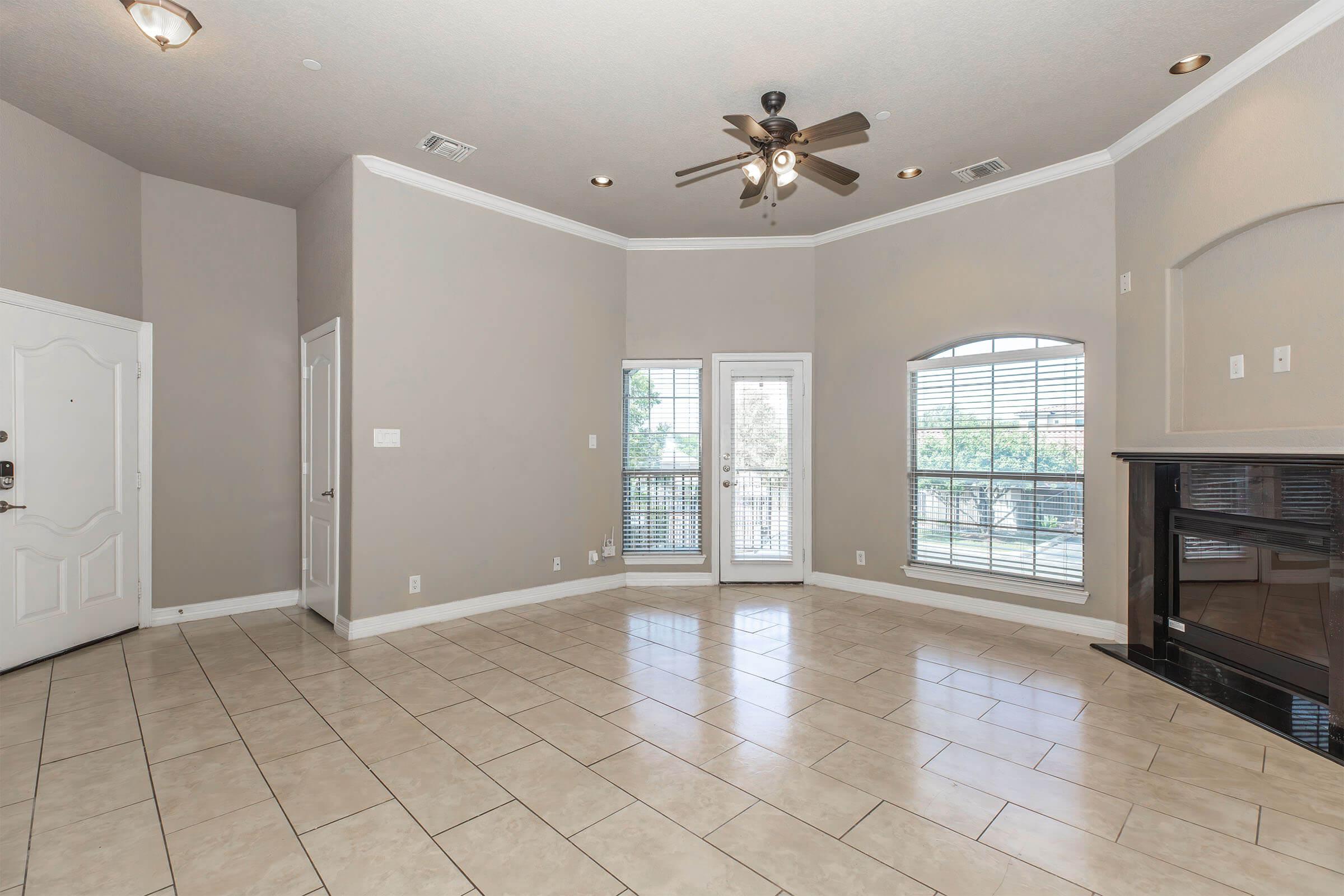 A spacious living room featuring tile flooring, neutral-colored walls, a ceiling fan, and large windows with shutters. There is a decorative fireplace on one side and a doorway leading to a balcony, enhancing natural light and openness.