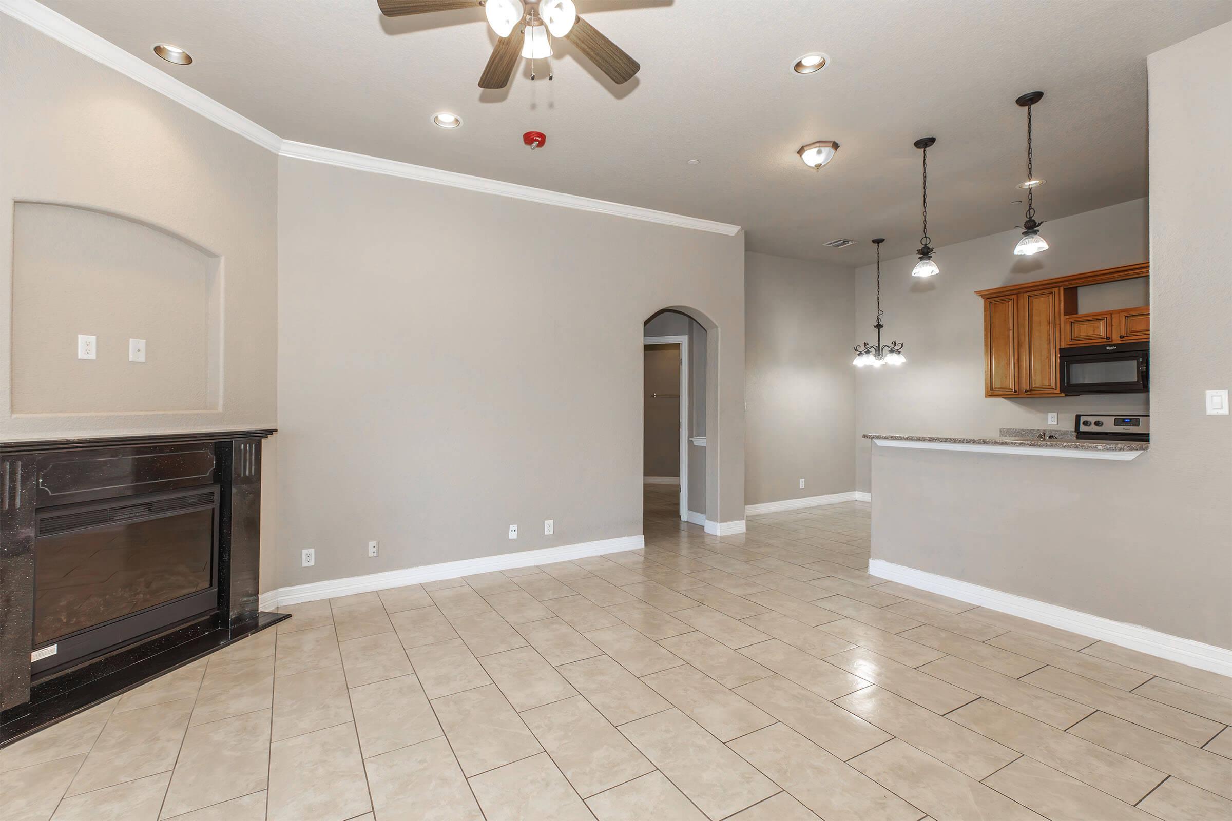 Spacious living room with tiled flooring, featuring a ceiling fan and archway leading to the kitchen area. The room has a decorative fireplace and is well-lit by recessed lighting. The adjoining kitchen includes wooden cabinetry and modern appliances.