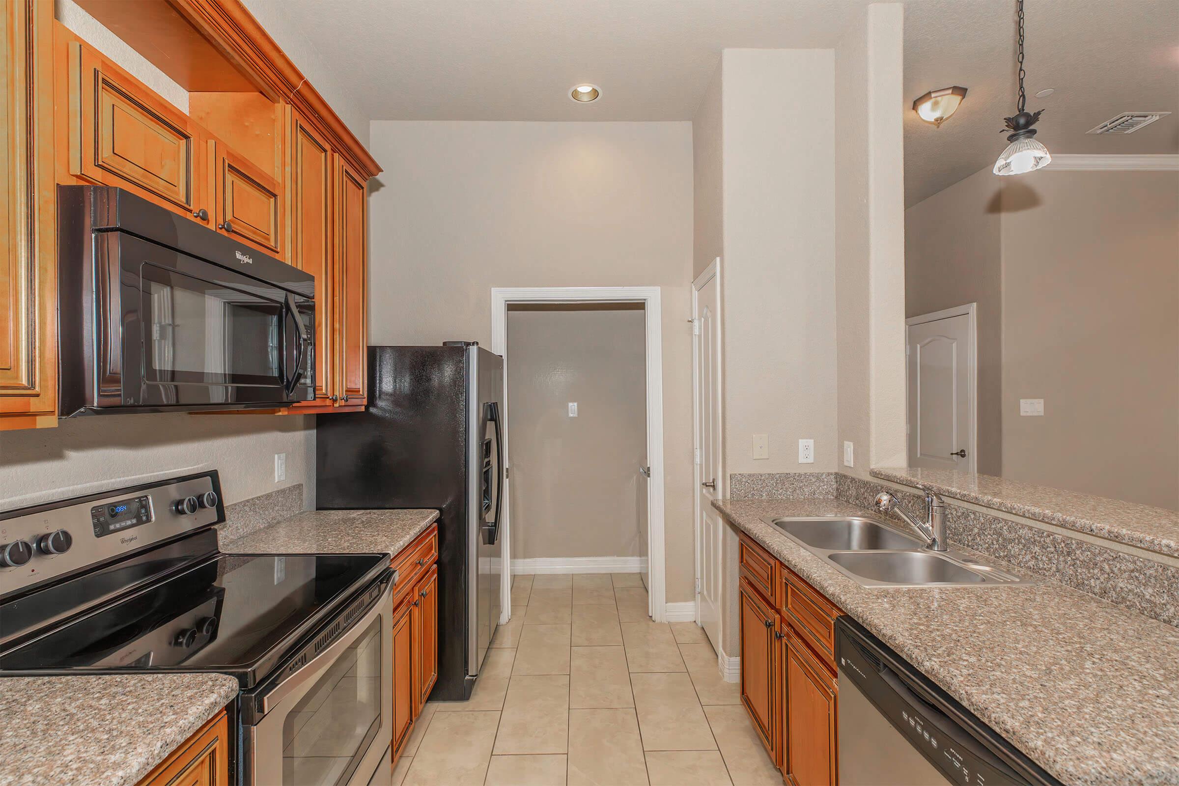 Modern kitchen featuring wooden cabinets, granite countertops, stainless steel appliances including an oven, microwave, and a double sink. Light-colored walls and tile flooring create a spacious feel. A doorway leads to an adjacent room, and overhead lighting adds a warm ambiance.