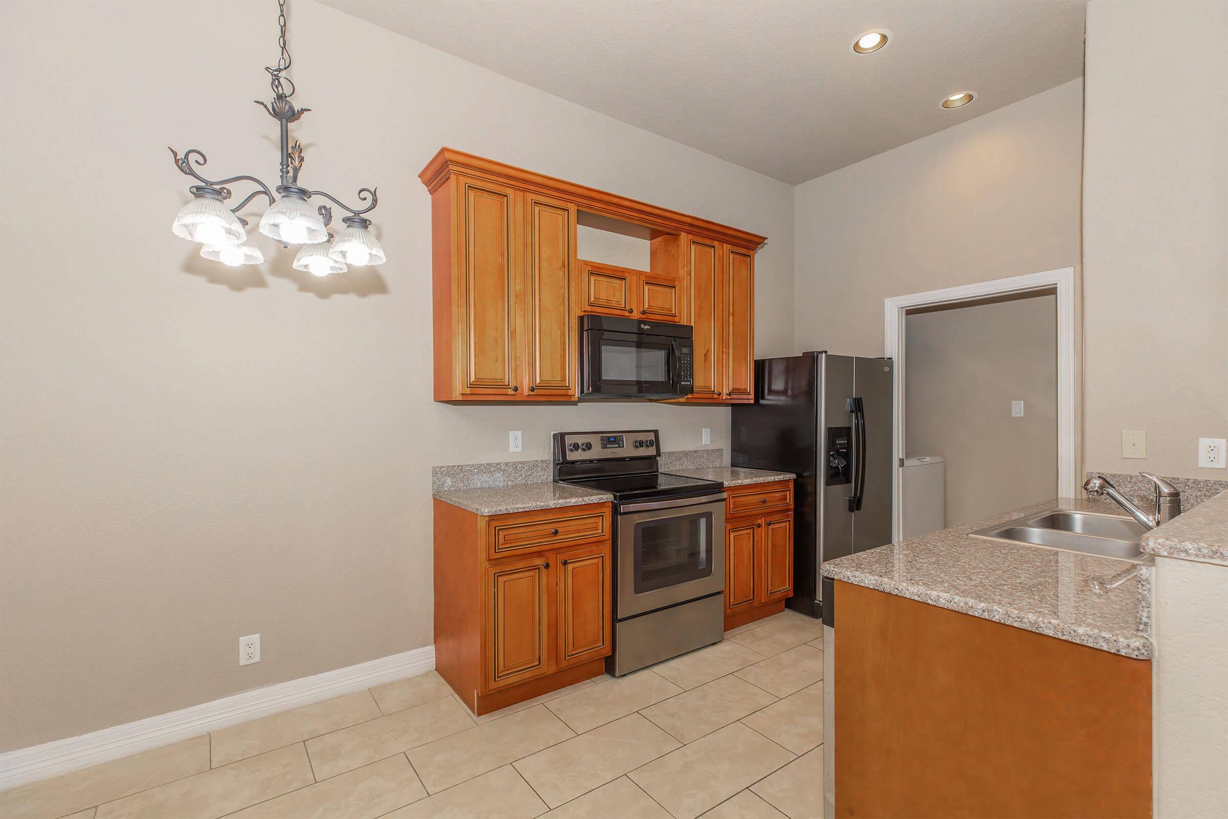 Modern kitchen featuring wooden cabinets, stainless steel appliances including a microwave and oven, and granite countertops. A chandelier hangs above the dining space. Neutral colored walls complement the tile flooring, providing a spacious and inviting atmosphere.
