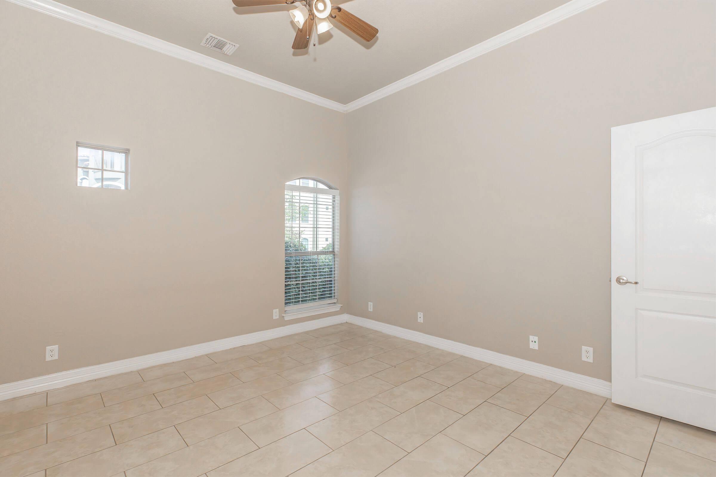 Empty room with light beige walls, tile flooring, and a ceiling fan. A small window is on the left wall, while a larger arched window with white blinds is on the right. The door is white with a silver handle, and there are no furnishings in the space, creating a bright and open atmosphere.