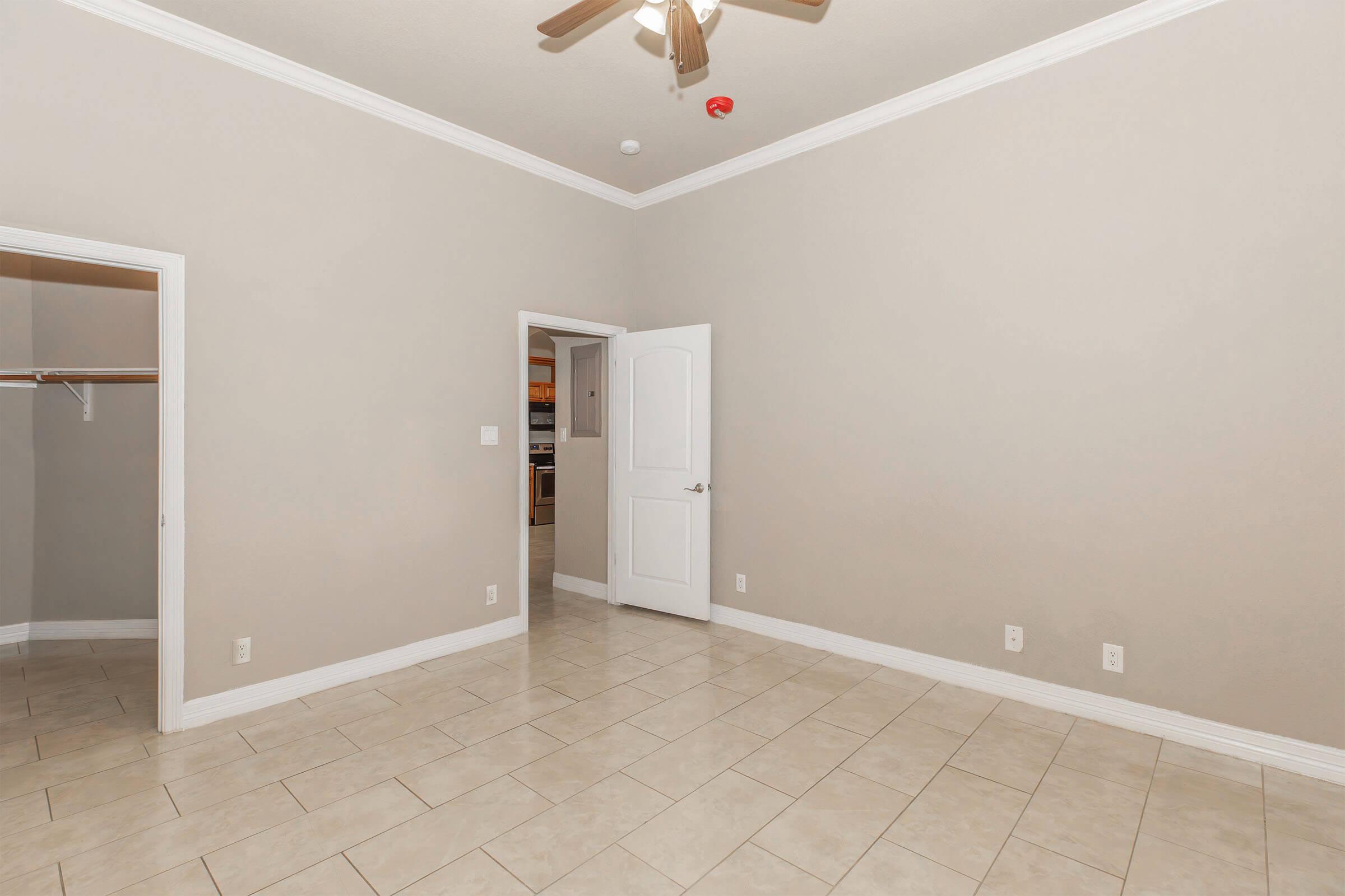 A spacious, empty room with light beige walls and tile flooring. There's a ceiling fan, a door leading to another room, and a closet on the left. The overall ambiance is bright and neutral, suitable for various decorating styles.