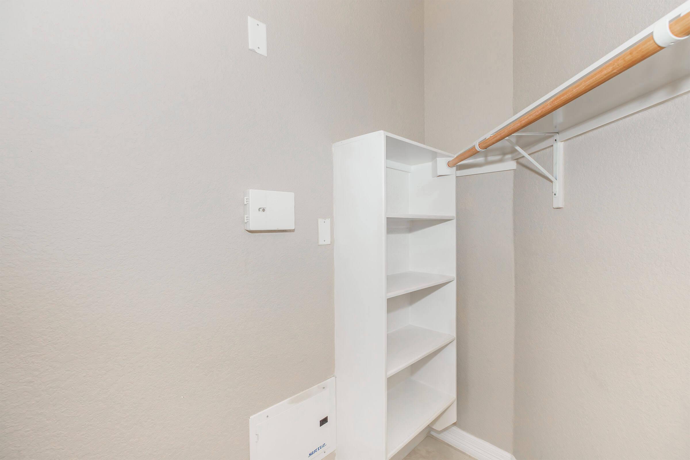 A well-organized closet featuring a white shelving unit with four open shelves, a clothing rod above, and light gray walls. There's a small electrical panel to the left and the floor is plain, making it a clean and functional space for storage or clothes.