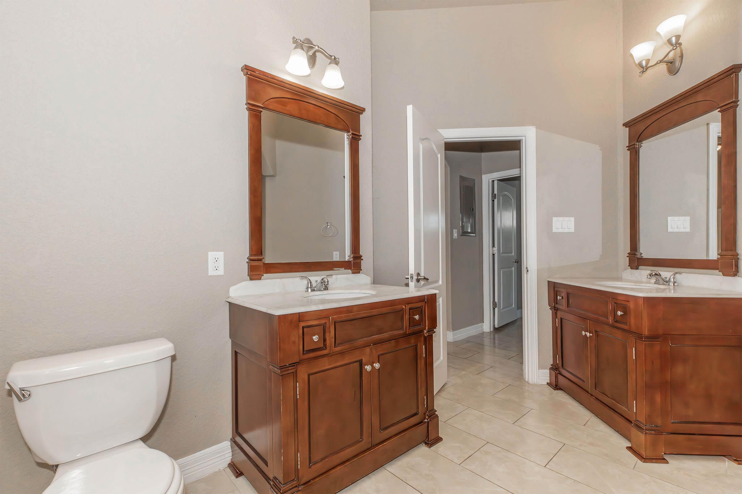 A spacious bathroom featuring two wooden vanities with sinks, large mirrors above each vanity, a white toilet, and neutral-colored walls. The tile floor complements the decor, and there are two light fixtures on the wall. A door leads to another room.