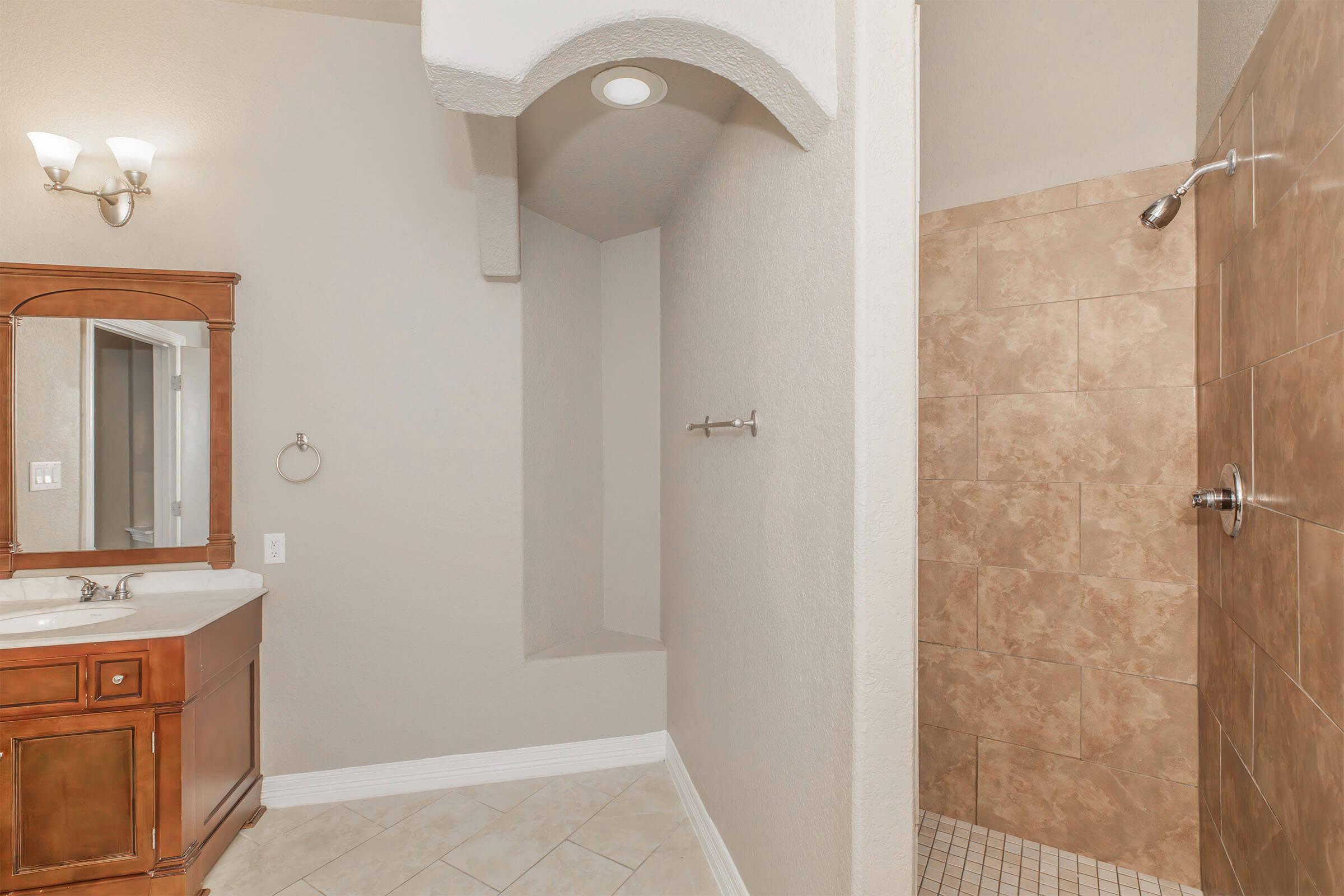 A bathroom featuring a spacious shower with tiled walls, a small built-in bench, and a vanity with a wooden cabinet and mirror. The walls are painted in a light color, and there are modern light fixtures. The floor is covered with large tiles, creating a clean and contemporary look.