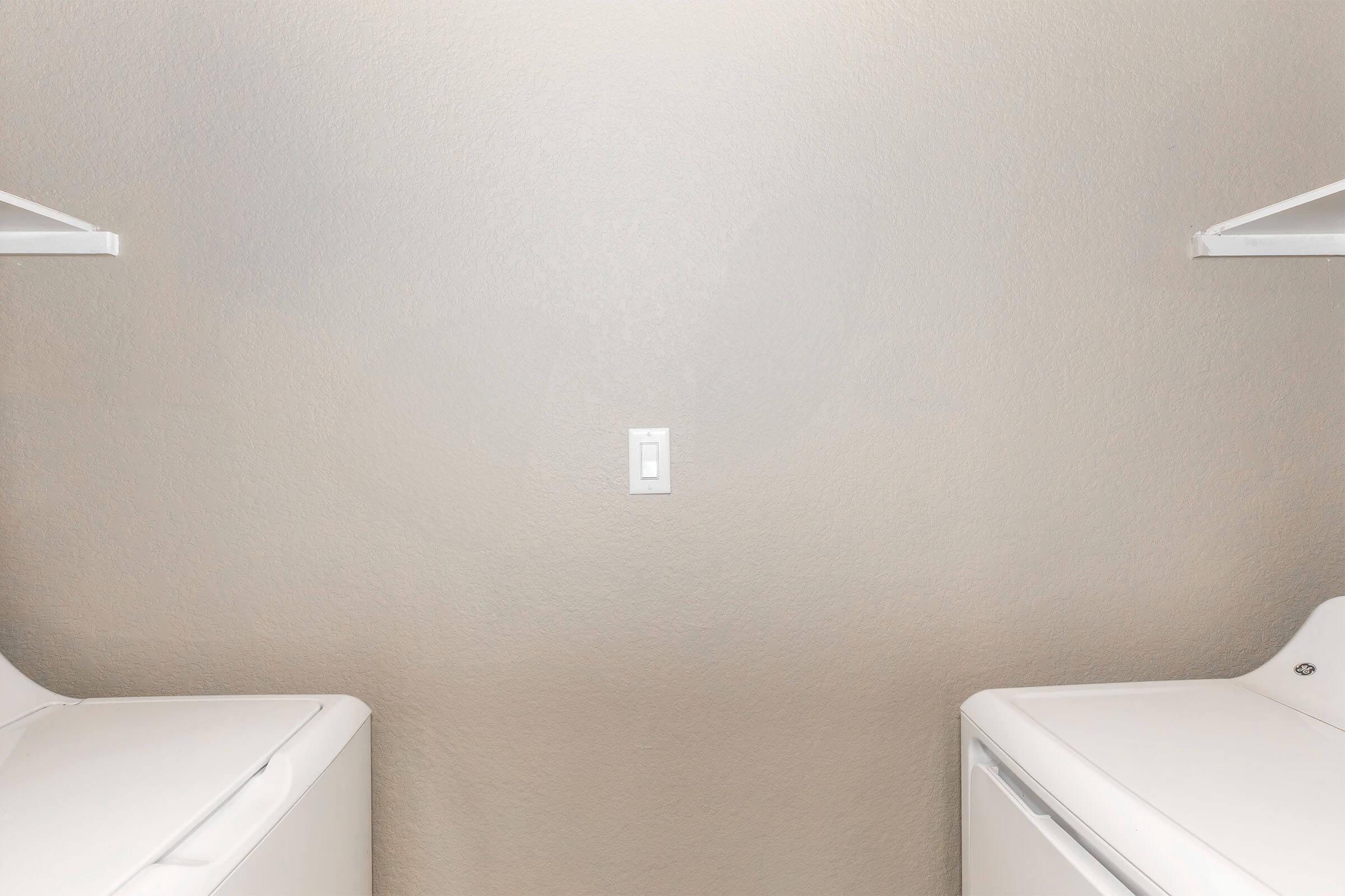 A laundry room with two white washing machines positioned on either side, against a plain beige wall. A light switch is centrally located on the wall, above a clean, unobtrusive floor. The overall atmosphere is clean and minimalistic.