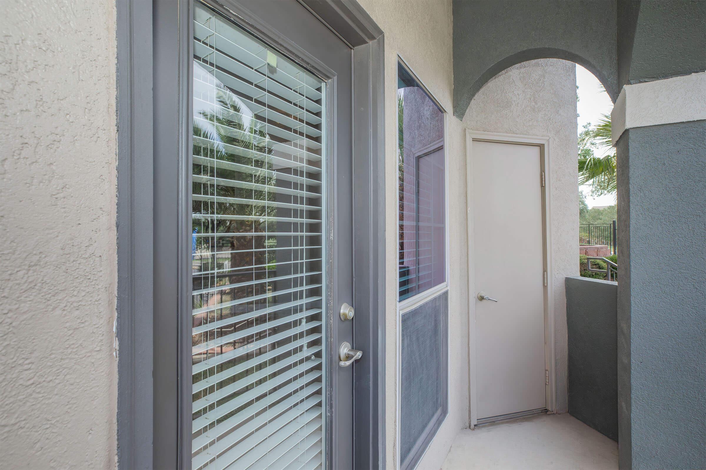A door with horizontal blinds, leading to a balcony area, is framed by a stucco wall. Next to it, there's a second door partially open, set in a similarly styled archway, with a view of green foliage outside. The overall color scheme includes gray and light beige tones.