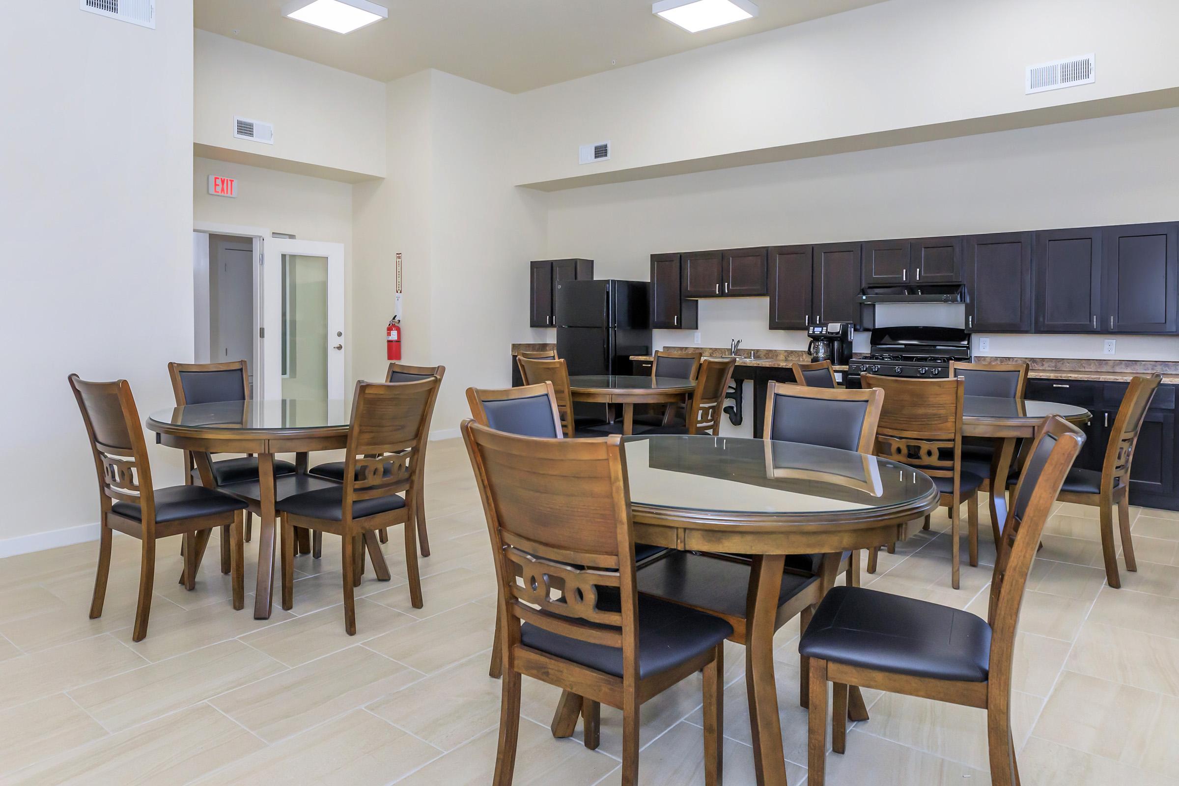 A spacious kitchen and dining area featuring several round tables with wooden chairs. The room has modern cabinetry, black appliances, and bright overhead lighting. Neutral-colored walls and tile flooring create a clean and welcoming atmosphere.