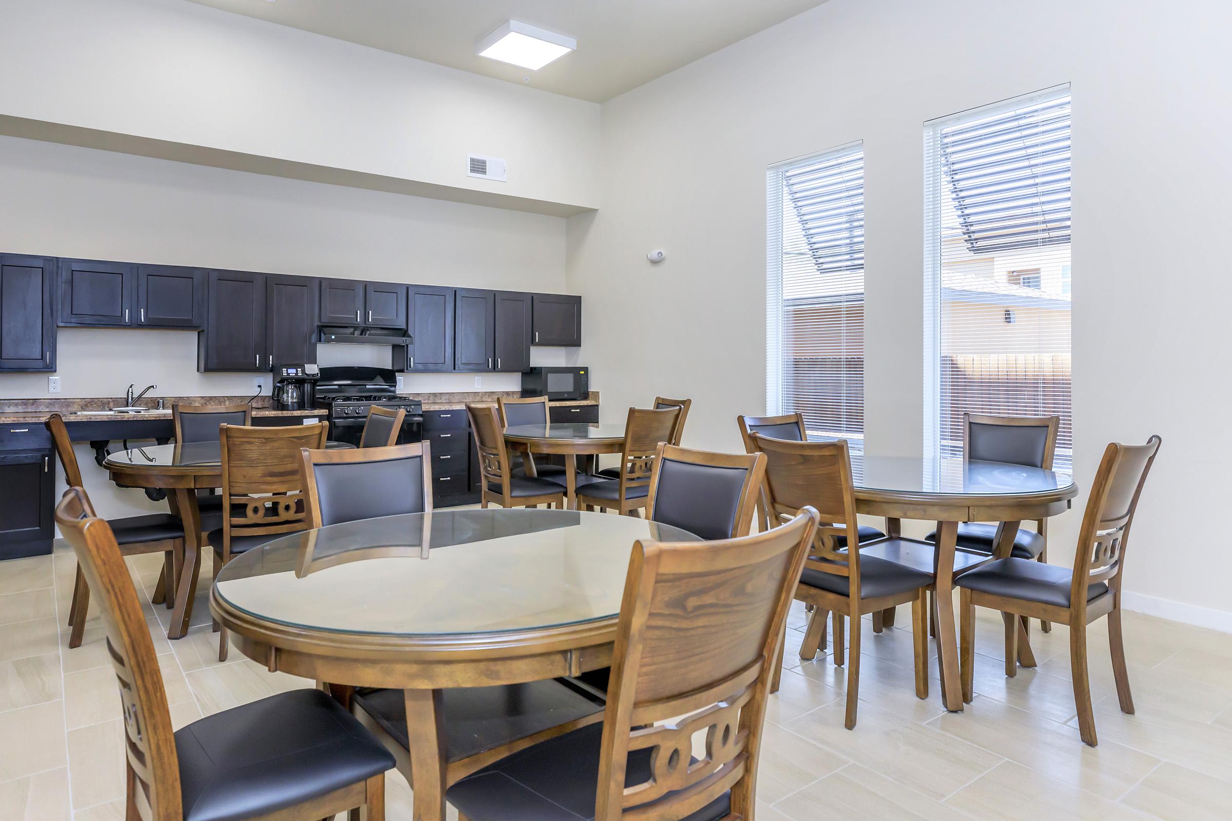 A spacious and modern dining area featuring multiple round wooden tables surrounded by black leather chairs. There is a well-equipped kitchen in the background with dark cabinetry, a stove, and microwaves. Large windows let in natural light, creating a welcoming atmosphere.
