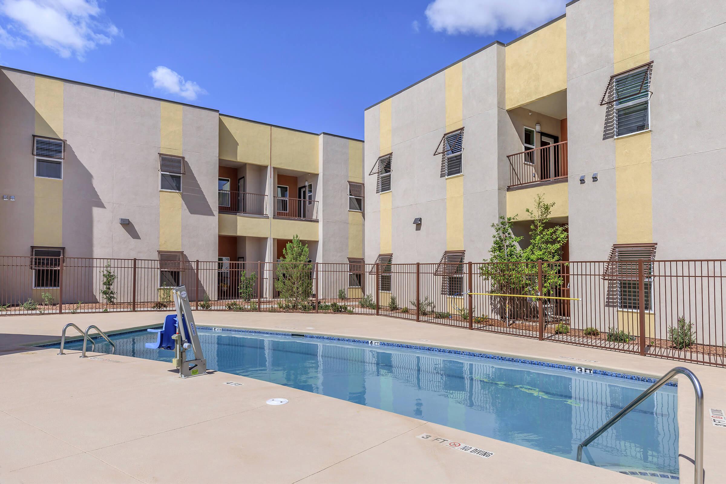 A well-lit courtyard with a swimming pool surrounded by modern apartment buildings. The pool features a ladder for entry, and there are potted plants and a blue slide next to it. The buildings have a blend of beige and yellow exterior with balconies. Clear blue skies are visible in the background.
