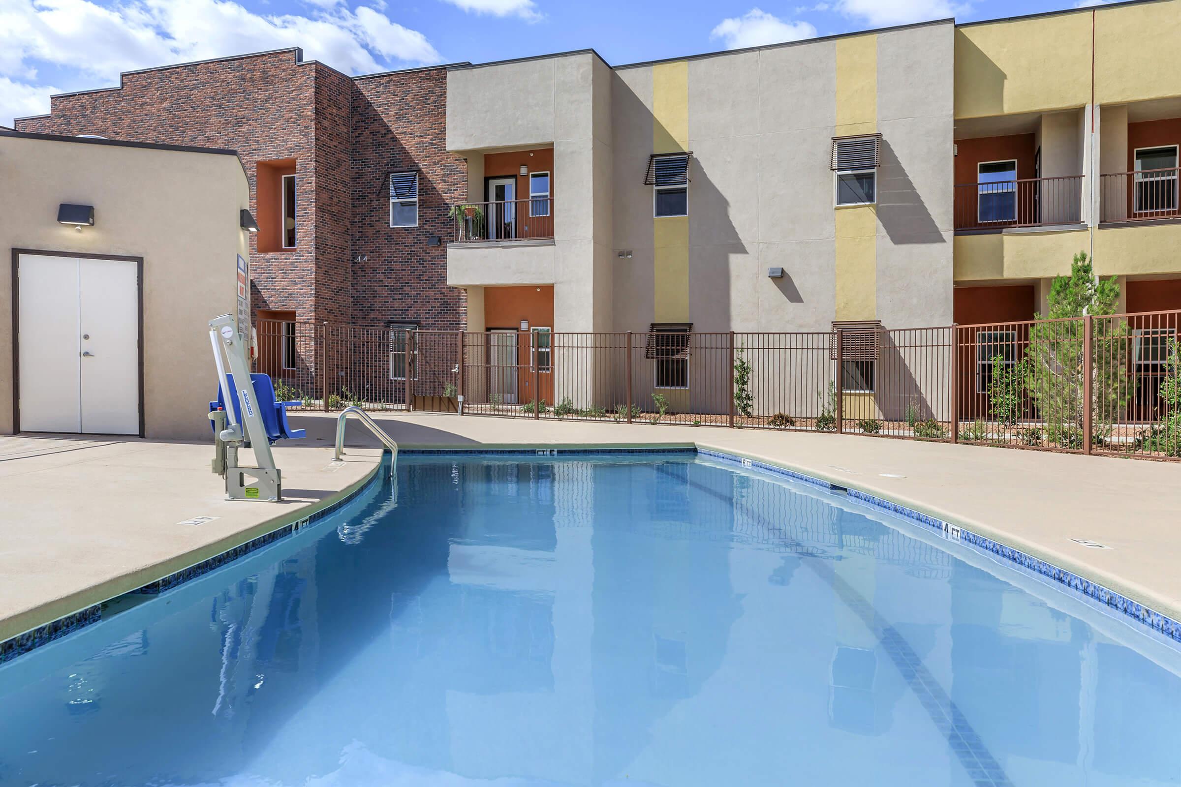 A clear swimming pool surrounded by a landscaped area and modern apartment buildings. The pool features a diving board and is bordered by a fence. The buildings have varying colors and architectural styles, with large windows and balconies. Bright blue sky with scattered clouds completes the scene.
