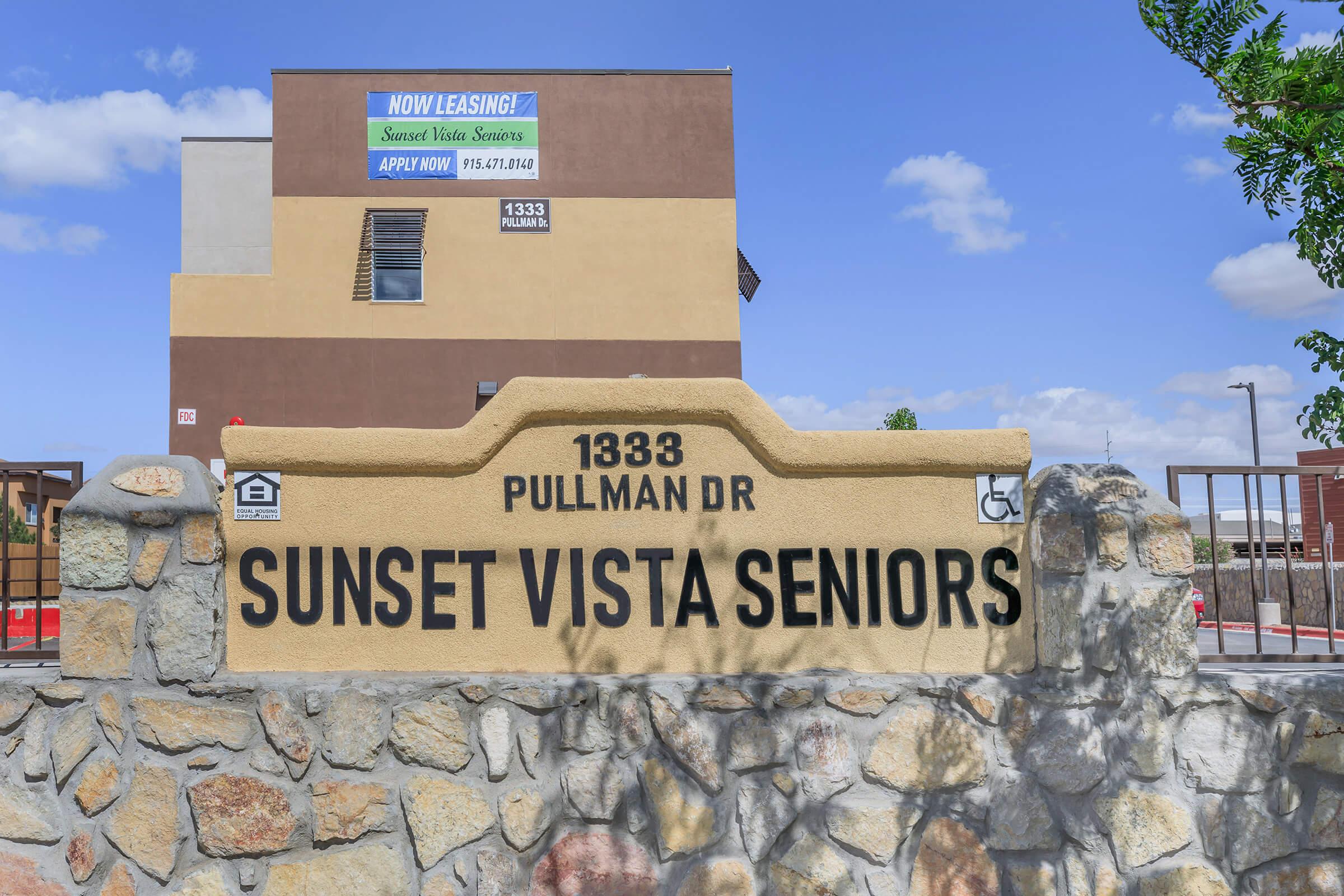Sign displaying "SUNSET VISTA SENIORS" on a stone wall, with the address "1333 PULLMAN DR." Above the sign, there is a banner that reads "NOW LEASING!" along with instructions to apply. The background features a building under a clear blue sky.