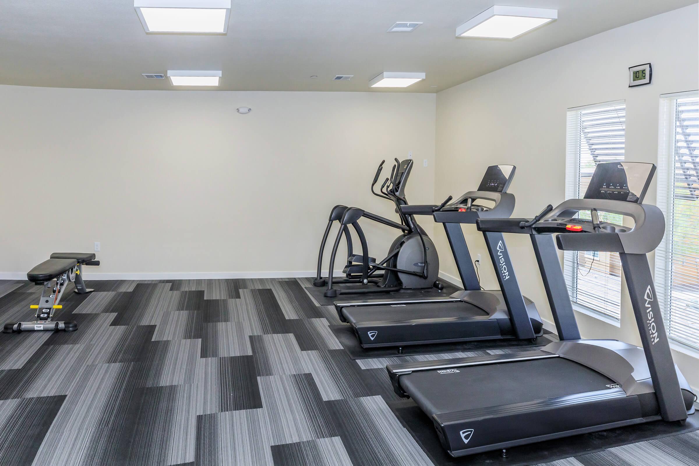 A modern gym space featuring two treadmills, an elliptical machine, and a weight bench. The room has neutral-colored walls and large windows with natural light, along with a patterned gray and black floor. Bright overhead lights enhance the indoor environment, making it suitable for workouts.