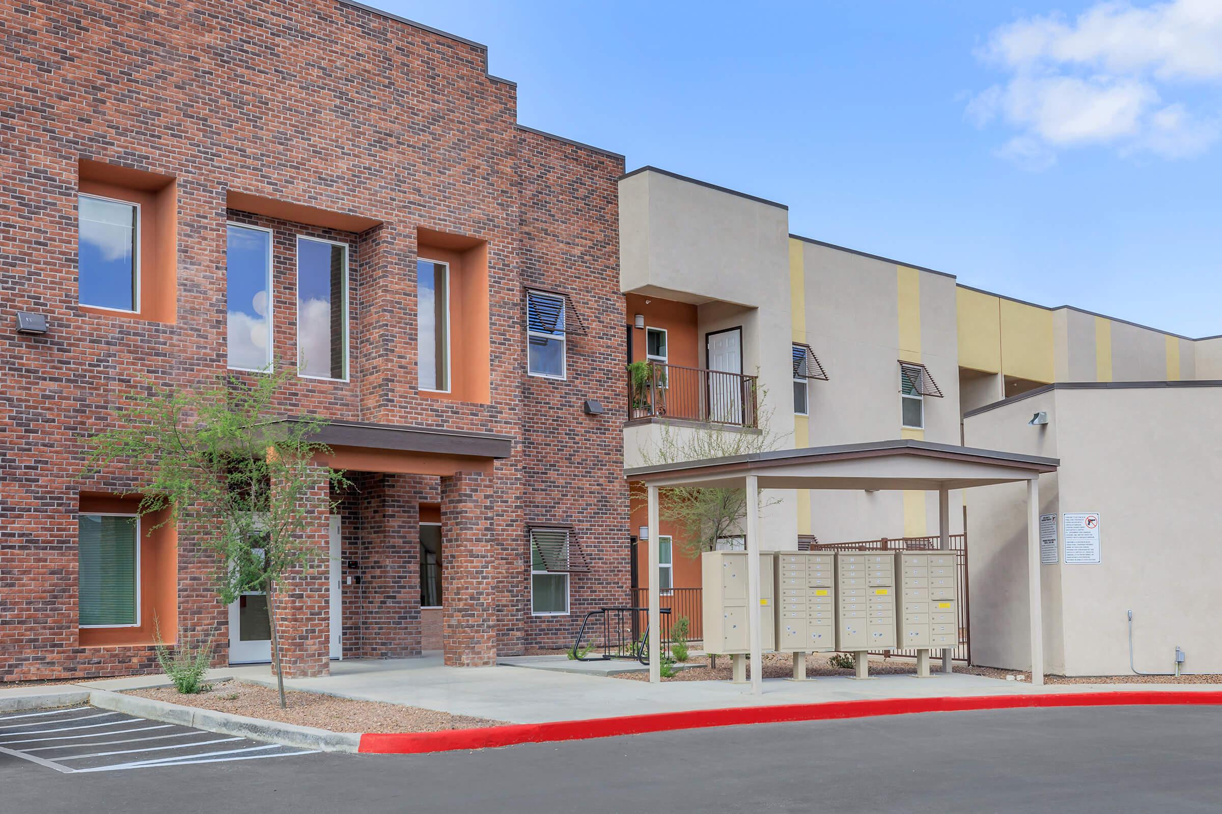 Modern apartment complex featuring a mix of brick and stucco architecture. The building has large windows, a covered area with mailboxes, and landscaping with small shrubs. Parking spaces are visible in front, and the sky is partly cloudy.
