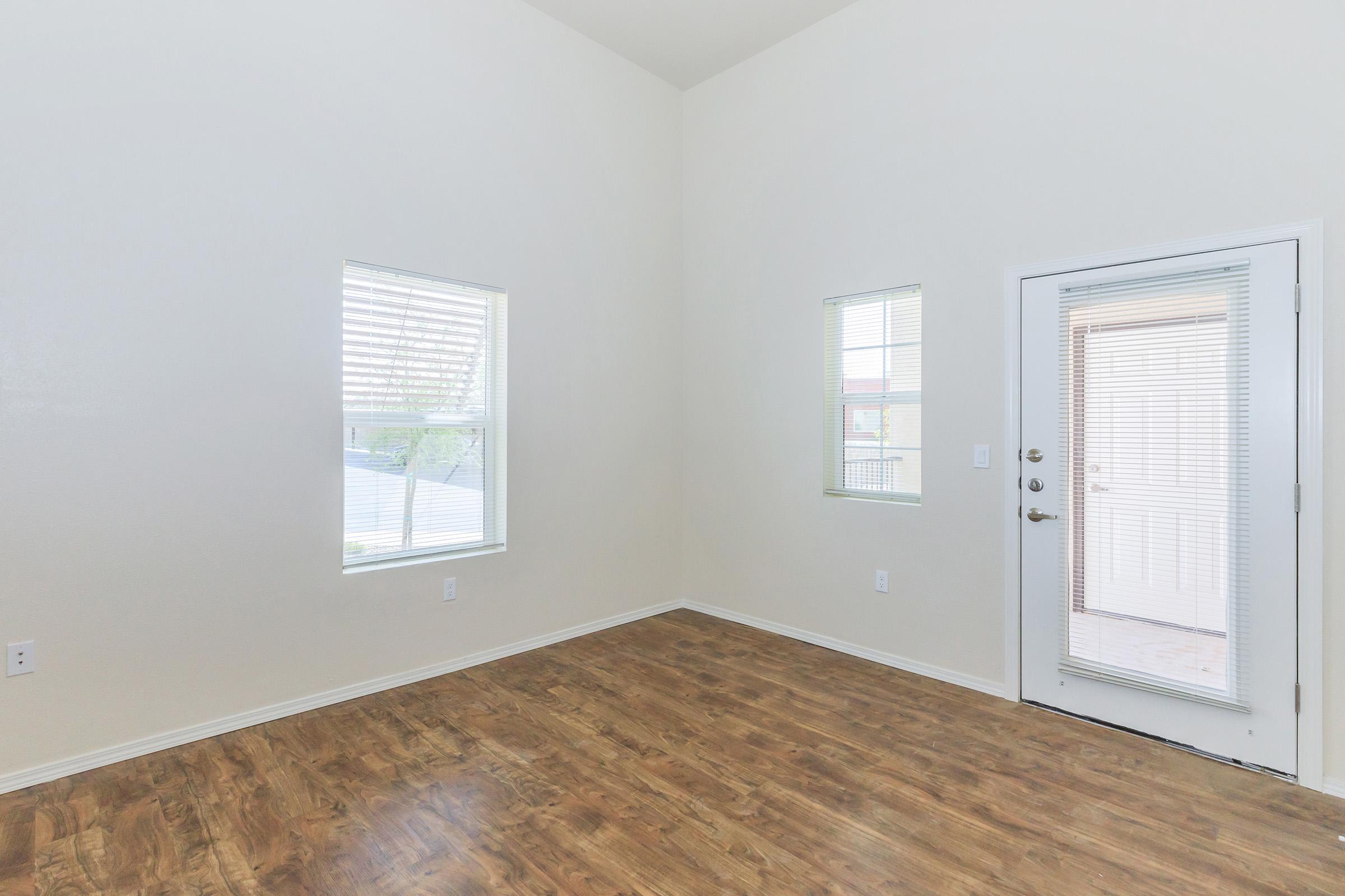 A bright, empty room featuring light-colored walls and a high ceiling. It has two windows letting in natural light, and a door leading outside. The floor is made of polished wood, creating a clean and open space ready for furnishing.