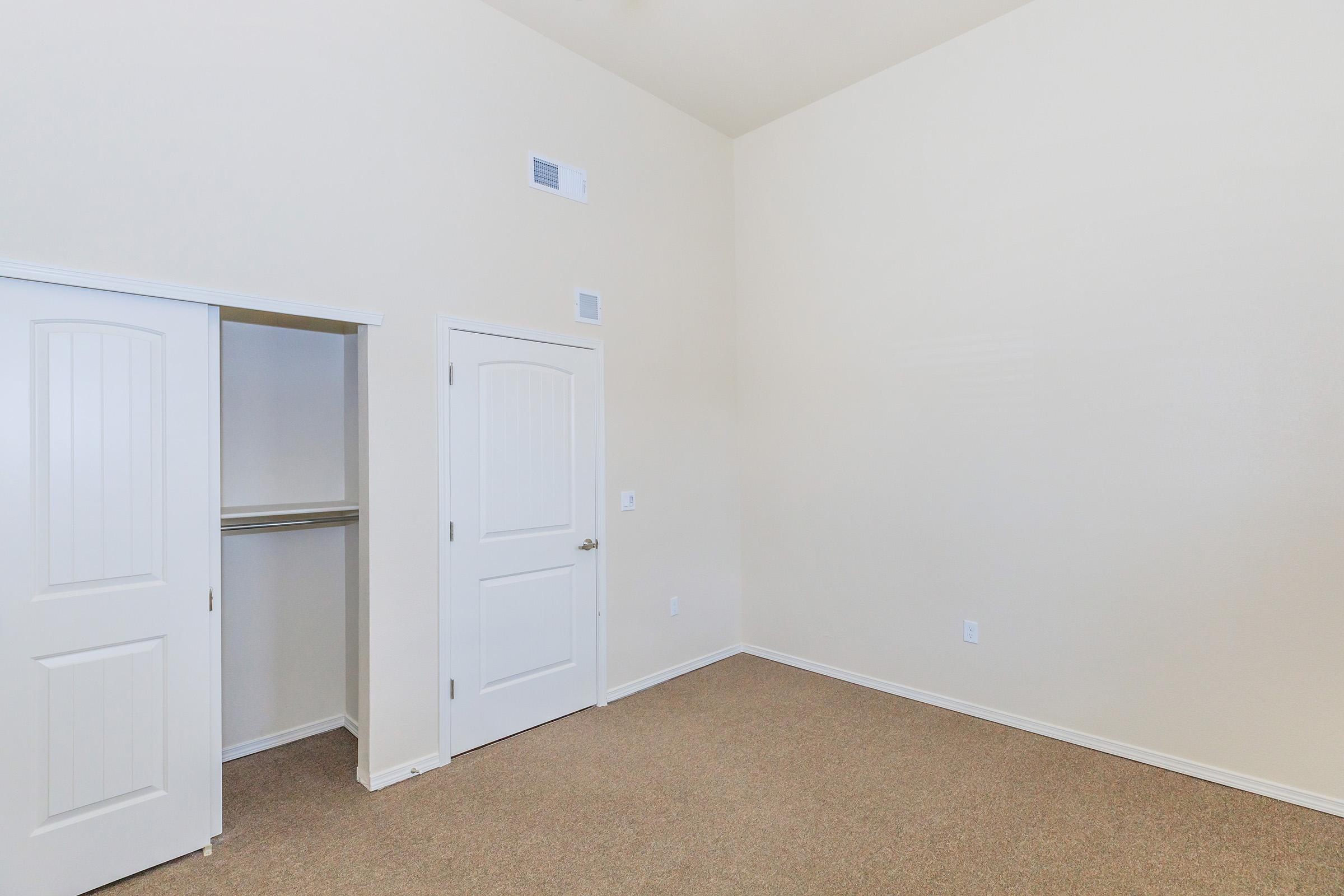 Empty room with light beige walls and carpet flooring. There is a white closet with double doors on the left and a single white door on the right. The ceiling is high, contributing to the spacious feel of the room, which has minimal furnishings or decorations.