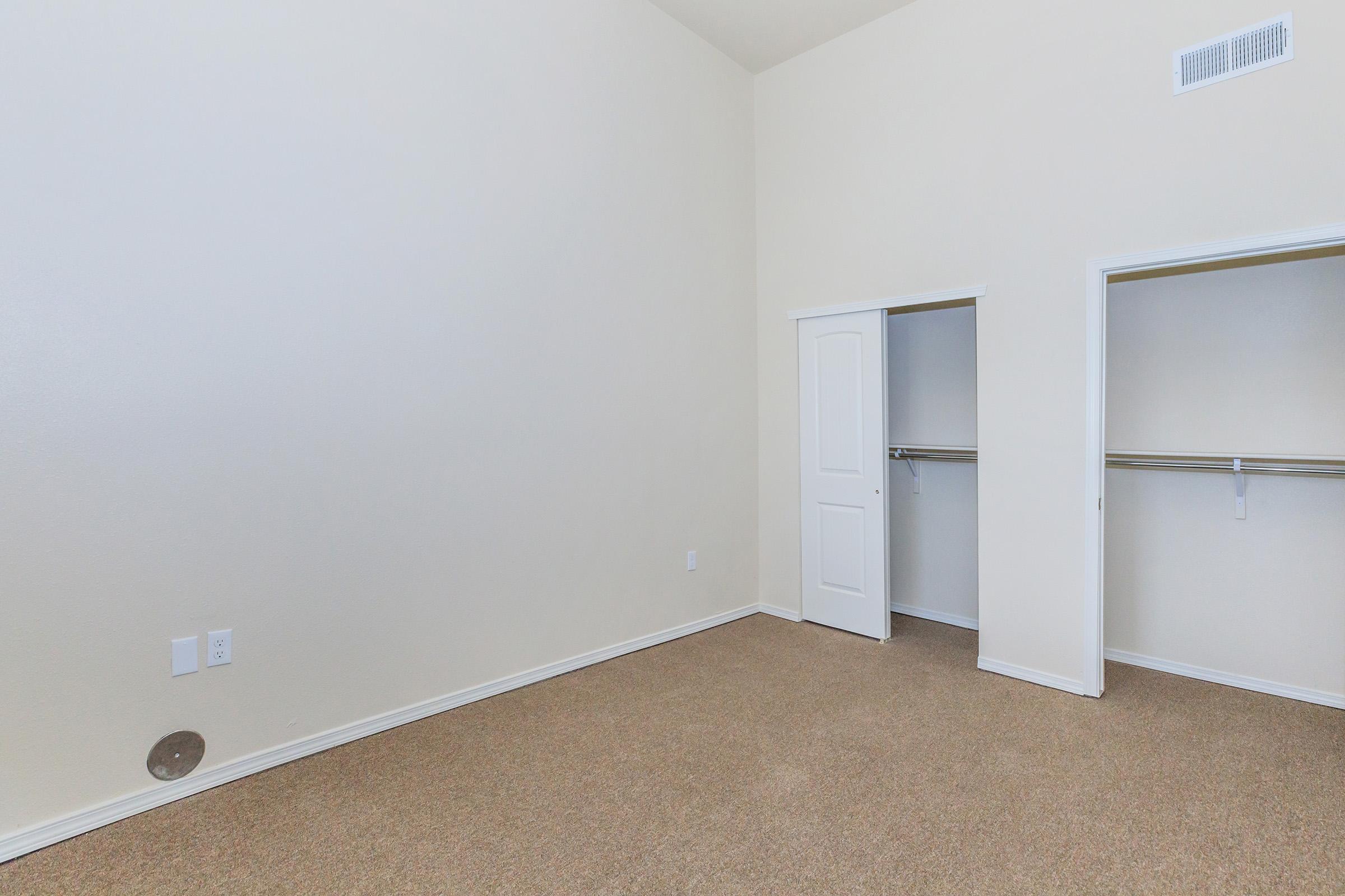 A spacious, empty room with beige carpet and light-colored walls. There are two open closet doors on the right side, showing hanging rods inside. The ceiling appears high, enhancing the sense of openness. A small round outlet is visible on the left wall.