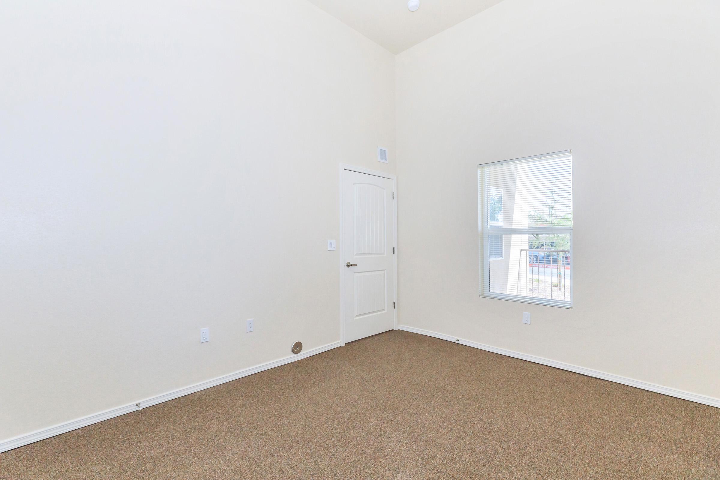 A spacious, empty room with beige walls and a high ceiling. There is a closed white door and a window with blinds allowing natural light. The floor is covered with brown carpet. The space is well-lit and inviting, suitable for various uses like a bedroom or office.