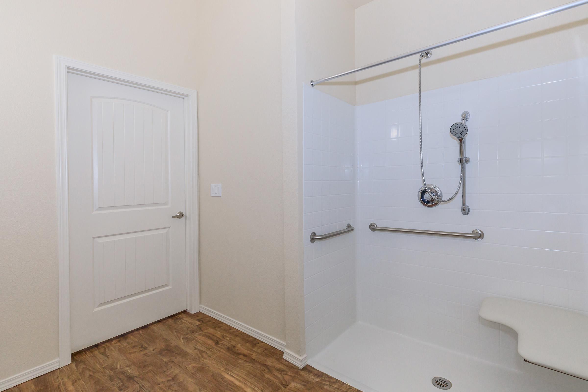 A bathroom featuring a walk-in shower with grab bars, a built-in bench, and a handheld showerhead. The walls are tiled in white, and the floor is made of wood-like laminate. A closed door is visible on the left side, leading to another area of the bathroom.