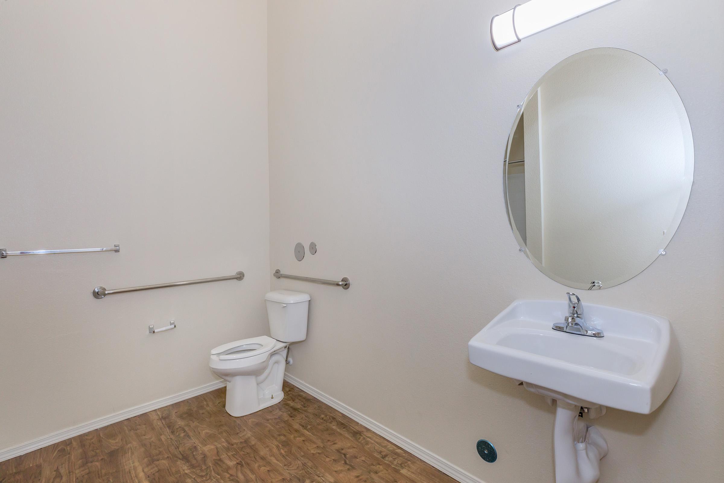 A clean, simple bathroom featuring a toilet, a wall-mounted sink, and a round mirror. The walls are painted a light color, and there are grab bars installed next to the toilet and sink. The floor has a wooden texture, contributing to a modern and minimalistic atmosphere.