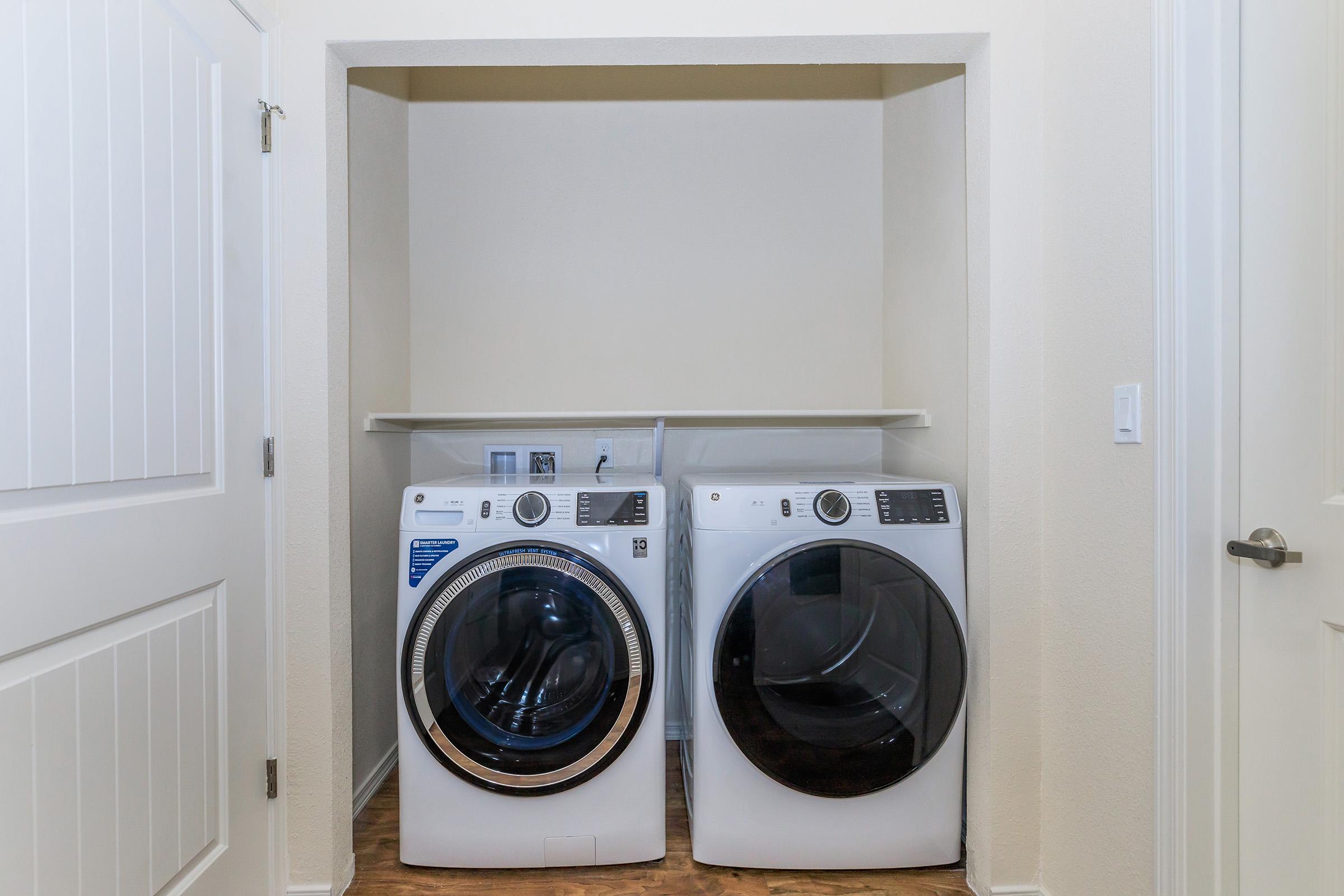 A laundry nook featuring two front-loading washing machines side by side, with a shelf above. The walls are painted in a light color, and a door is partially visible to the left. The flooring appears to be wood laminate.