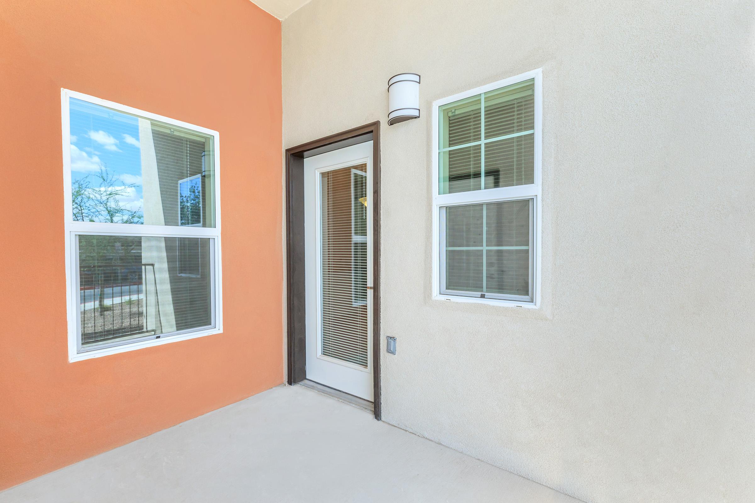 Exterior view of a modern entrance featuring a white door with glass panels, flanked by two windows. One wall is painted orange, contrasting with the light beige stucco of the other wall. The area appears well-lit and clean, with a minimalistic design and a contemporary feel.