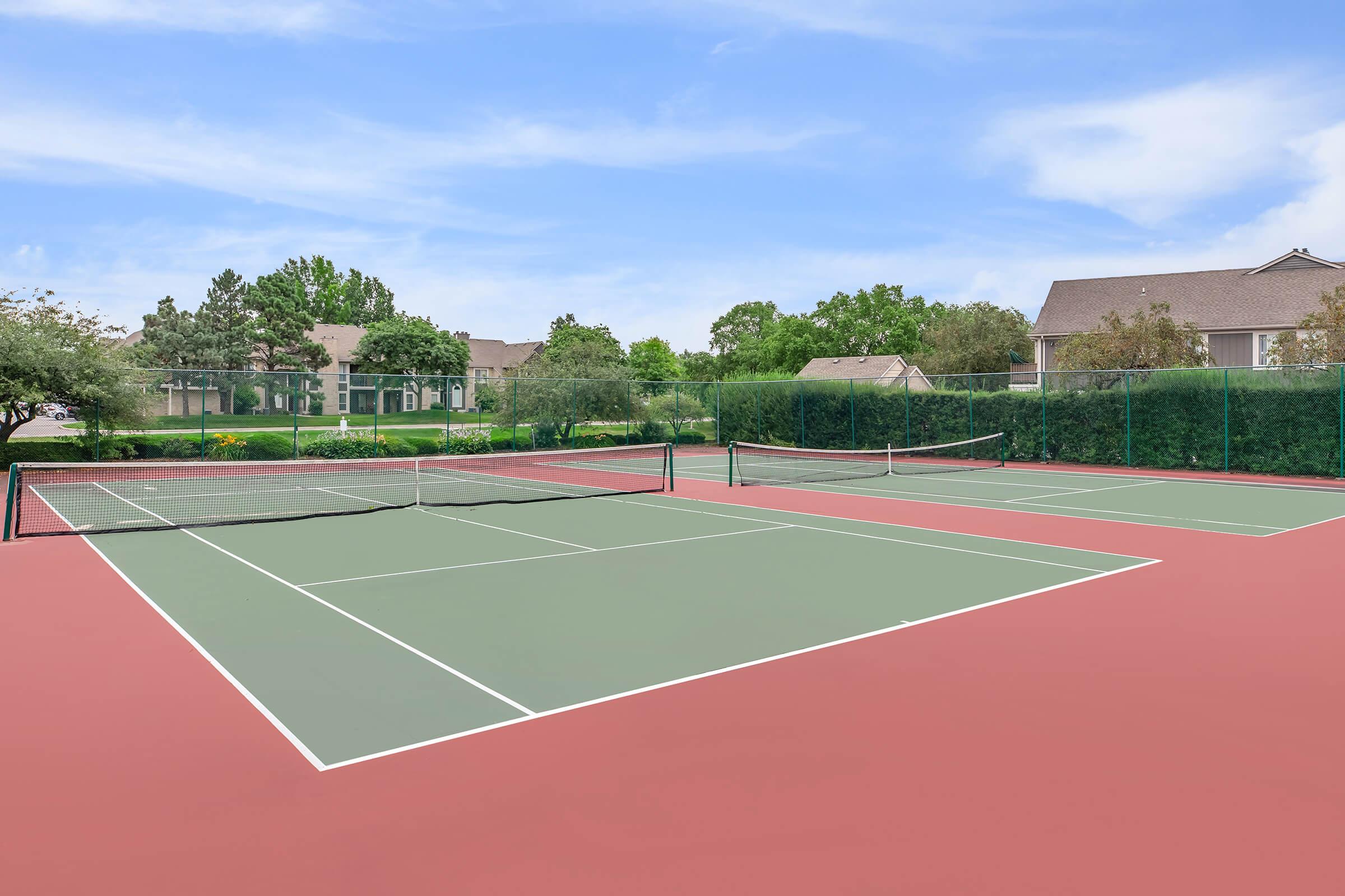 A view of two empty tennis courts surrounded by greenery, with a clear blue sky above. The courts feature red and green surfaces, and are enclosed by a fence. Nearby residential buildings and landscaped gardens are visible in the background.