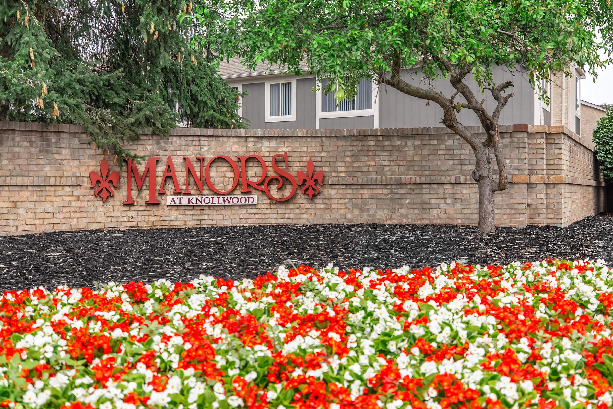 A brick sign reading "Manors at Knollwood" surrounded by vibrant red and white flowers. Lush green trees and a well-maintained landscape enhance the inviting atmosphere of the property entrance.