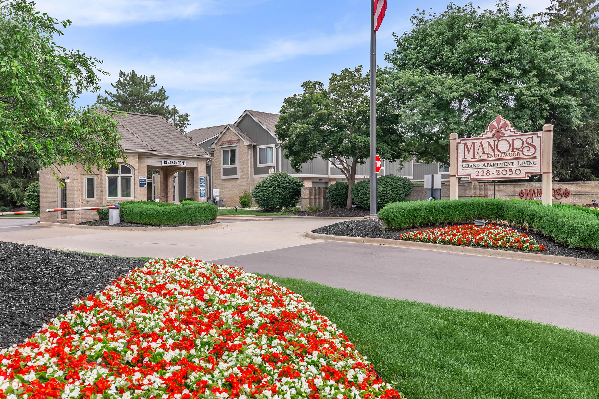 A brick building with a sign reading "Manors Grand Apartment Living" is visible at the entrance. The scene features well-maintained landscaping with vibrant red and white flowers in the foreground, green grass, and trees. An American flag is positioned nearby, and a vehicle entrance barrier is present.