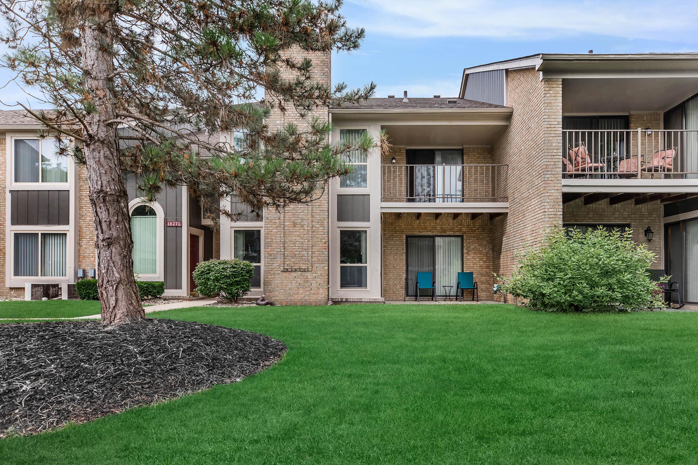 A two-story apartment building with brick exterior and a balcony. In the foreground, there is a well-manicured green lawn and a tree. The building features large windows, and two blue chairs are visible on the balcony. Mulch surrounds the base of the tree, enhancing the landscape.