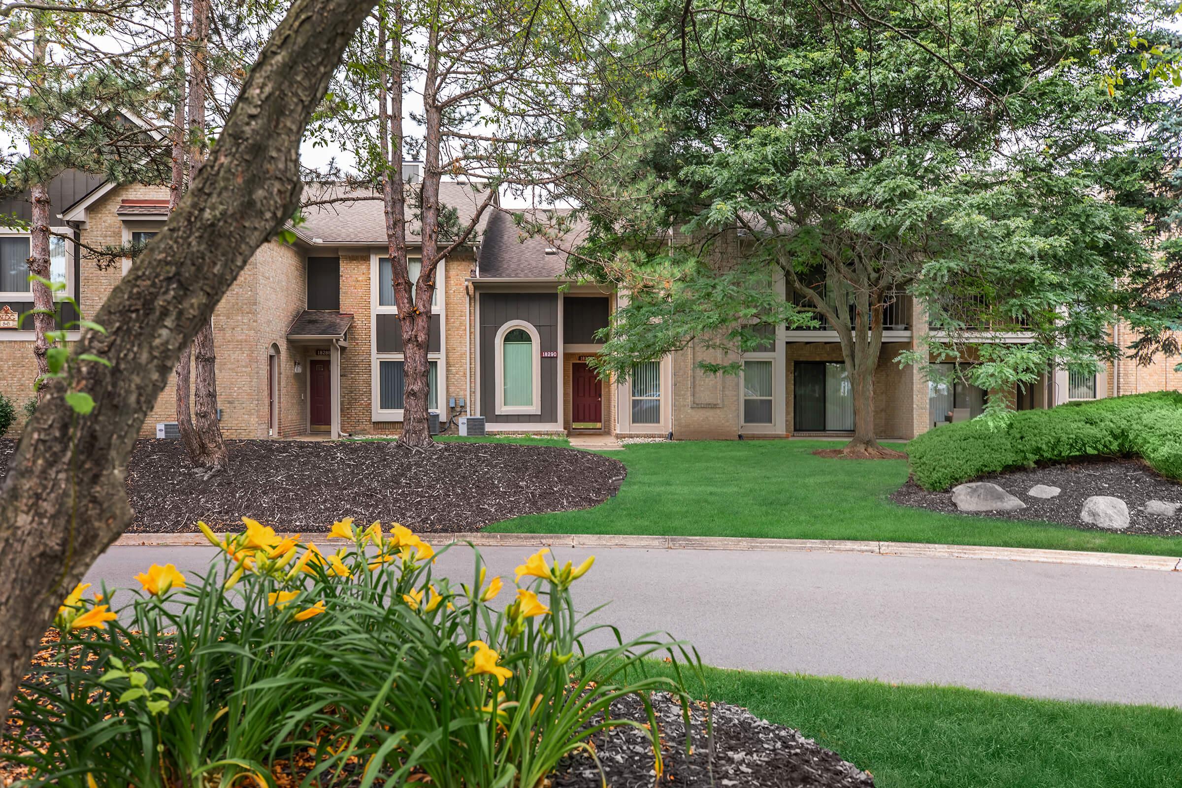 A landscaped area featuring vibrant yellow flowers in the foreground, with a well-maintained lawn and shrubs. In the background, there are multi-story townhouses with large windows and a mix of brick and siding exteriors, set among trees. The scene is peaceful and inviting, showcasing a residential neighborhood.