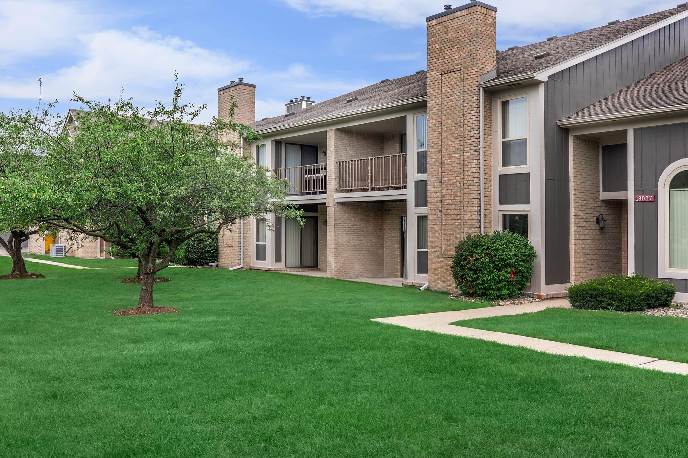 A view of a well-maintained apartment community with brick exteriors. The foreground features a vibrant green lawn and a small tree, while the two-story building has balconies and multiple windows. The sky is clear, adding to the pleasant atmosphere of the residential area.