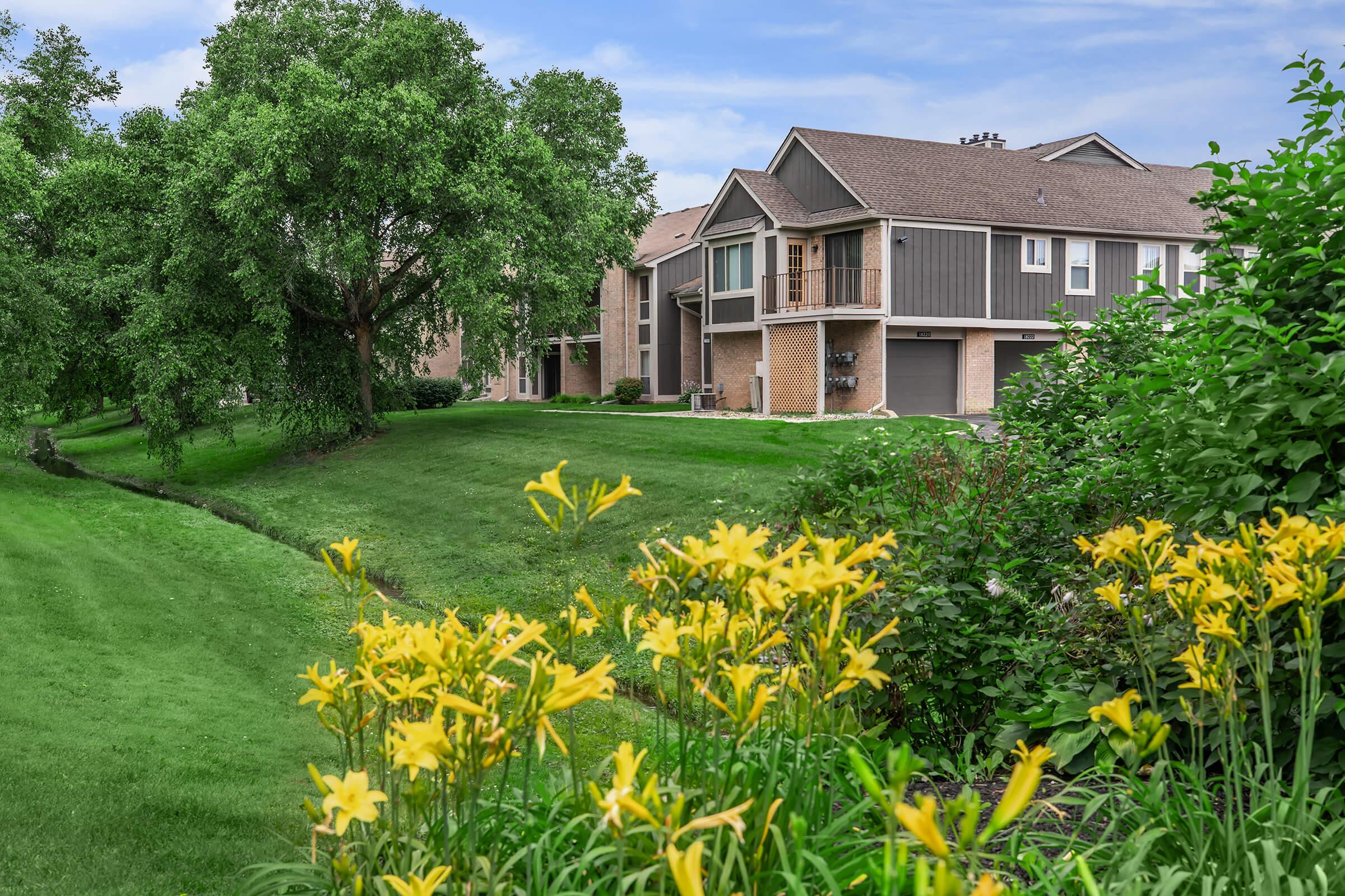 A serene residential scene featuring two houses bordered by lush green lawns and trees. In the foreground, bright yellow flowers bloom amid vibrant greenery, with a small stream or path visible on the left side, enhancing the peaceful suburban atmosphere.