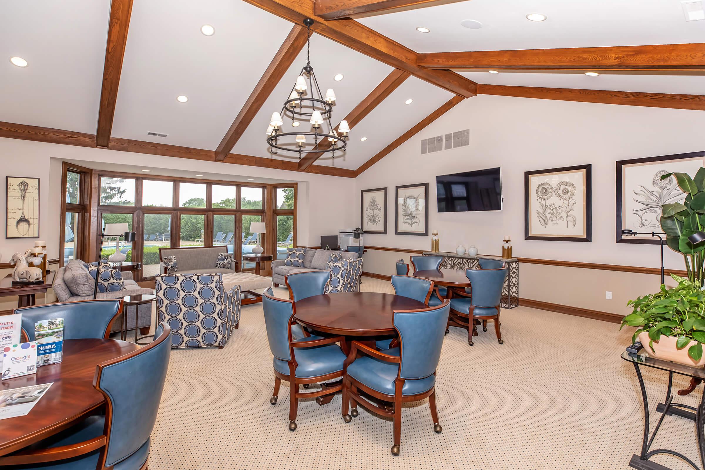 A cozy interior space featuring high wooden beams, elegant chandeliers, and plush seating. The room includes round dining tables with blue chairs, framed floral artwork on the walls, and large windows offering a view of the outdoors. A television is mounted on the wall, adding to the inviting atmosphere.