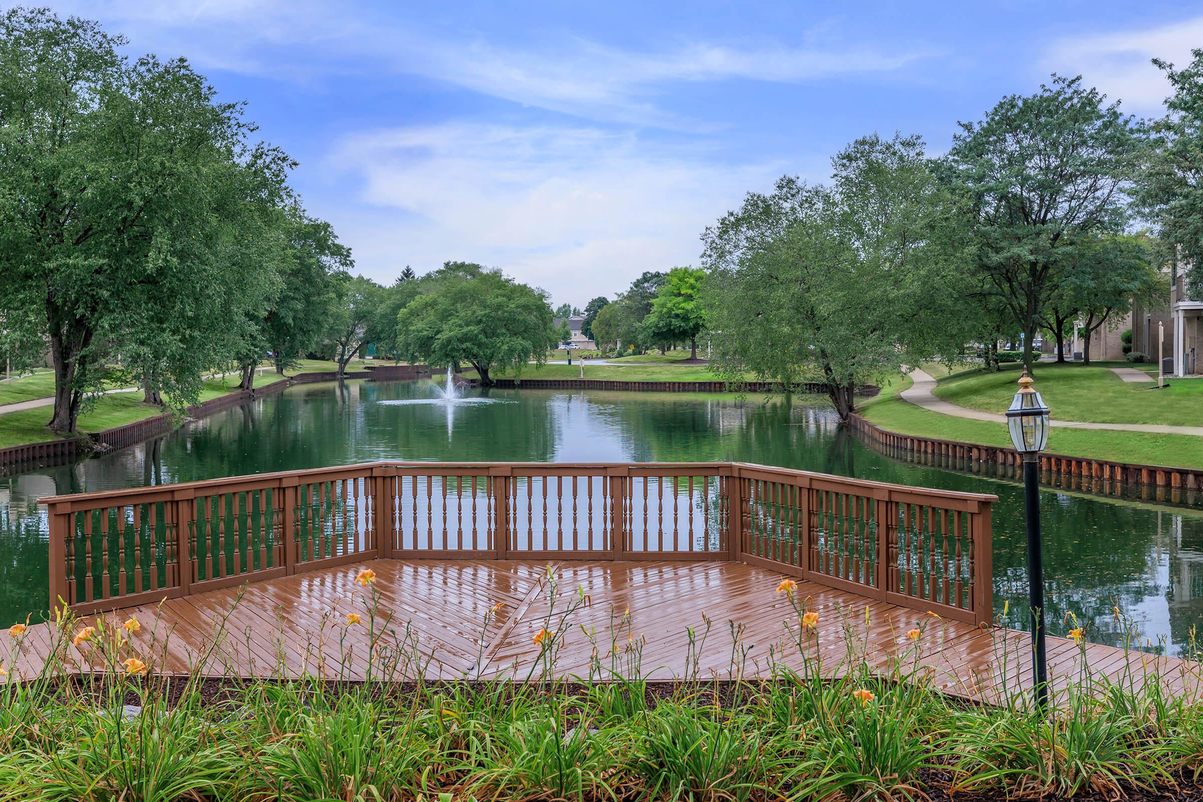 A tranquil scene featuring a wooden deck overlooking a serene pond surrounded by lush greenery and trees. The water reflects the blue sky and clouds, with a fountain gently spraying in the background. Flowering plants line the deck, enhancing the peaceful atmosphere of the setting.