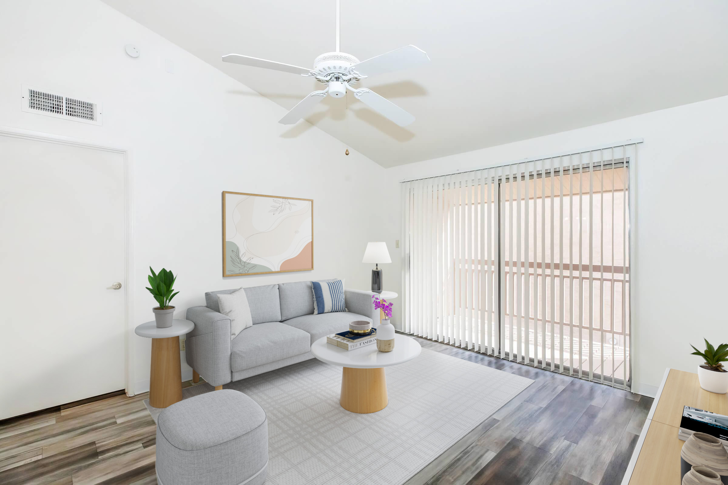 A bright and airy living room with a light gray sofa, a round coffee table, and a floor lamp. There are potted plants on the table and a cozy rug on the floor. Large windows with vertical blinds allow natural light to fill the space, creating a welcoming atmosphere. A piece of artwork is displayed on the wall.