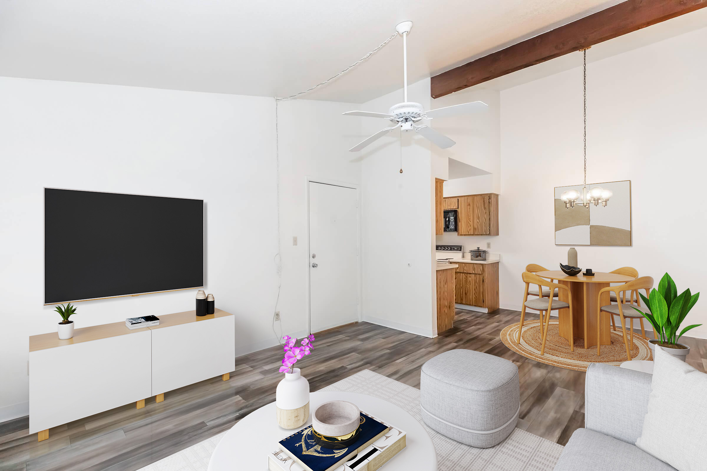 A modern, spacious living room featuring a wall-mounted flat-screen TV on a white media console, a cozy gray ottoman, and a coffee table with books and decor. A ceiling fan hangs above, and a small dining area with a round table and chairs is visible in the background, along with a kitchen. Natural light floods the space.
