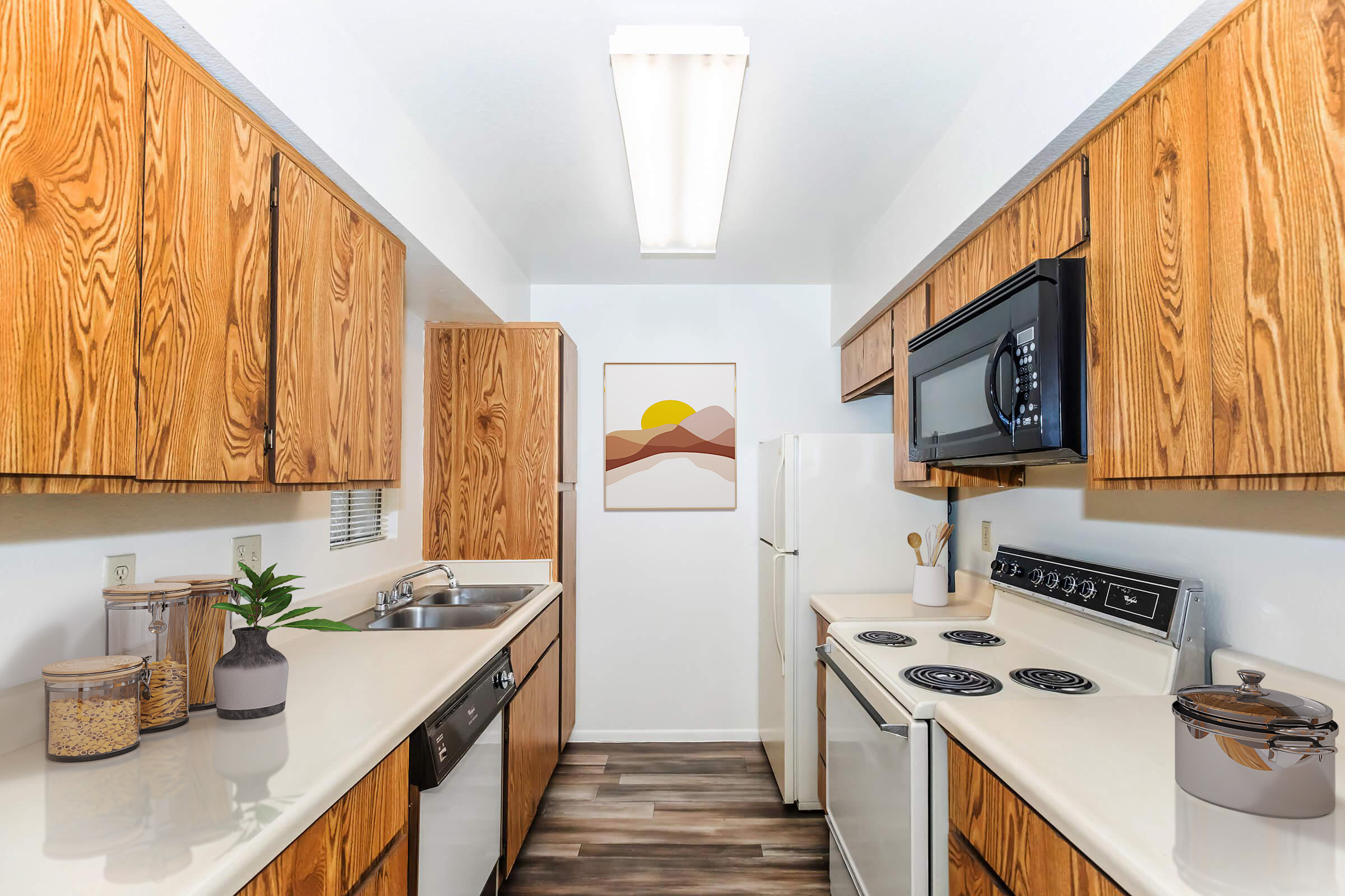 A cozy kitchen with wooden cabinets, a stainless steel microwave, and a white stove. It features a sink with a small plant and jars on the counter. A framed artwork with abstract mountains and a sun hangs on the wall, and the floor is covered in dark vinyl planks. The room is well-lit by a ceiling fixture.