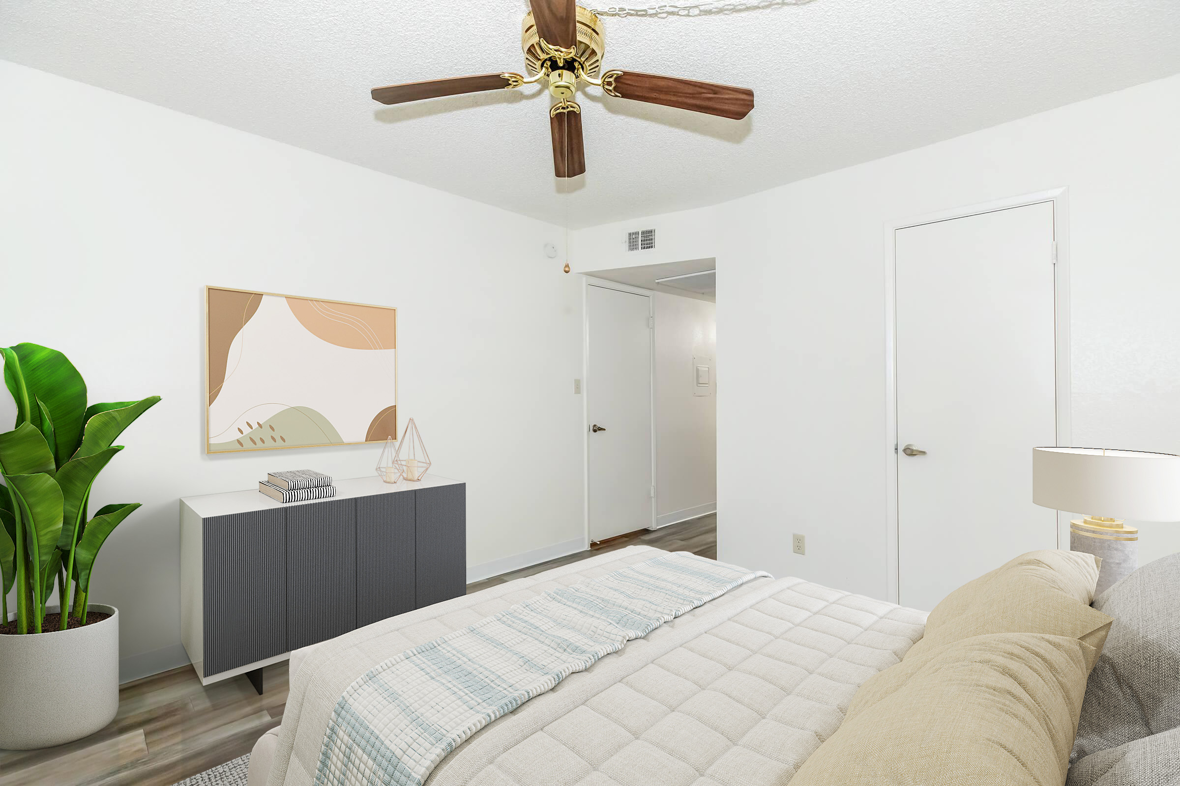 A well-decorated bedroom featuring a bed with neutral bedding, a ceiling fan, and a potted plant. A light-colored abstract painting is mounted on the wall above a modern dresser. The room has white walls and a door leading to another space. Natural light creates an inviting atmosphere.