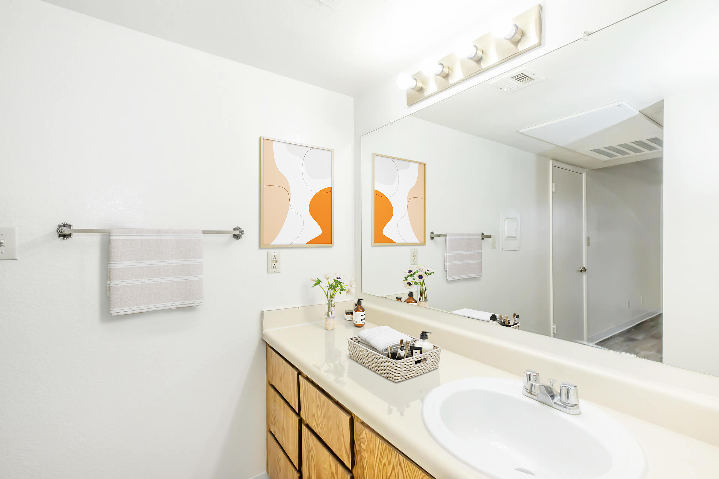 A modern bathroom featuring a light-colored vanity with a sink, a large mirror above it, and two abstract artworks on the wall. There are neatly arranged toiletries in a basket on the countertop, fresh flowers in small vases, and hand towels hanging by the sink. The space is well-lit and has a clean, minimalist design.