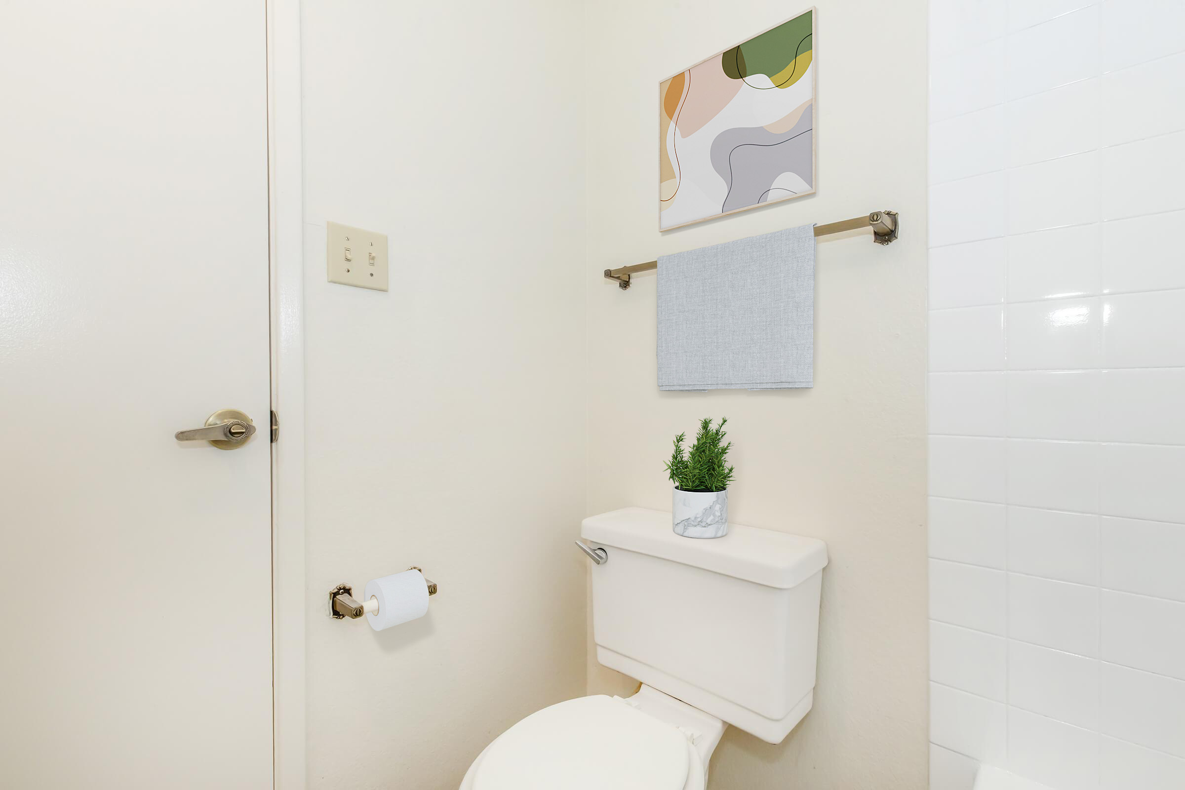 A clean bathroom corner featuring a white toilet and tank, a gray towel on a towel rack, a small potted plant on top of the toilet tank, and a framed abstract artwork on the wall. The decor is minimalist and modern, with neutral tones enhancing the bright, tidy atmosphere.