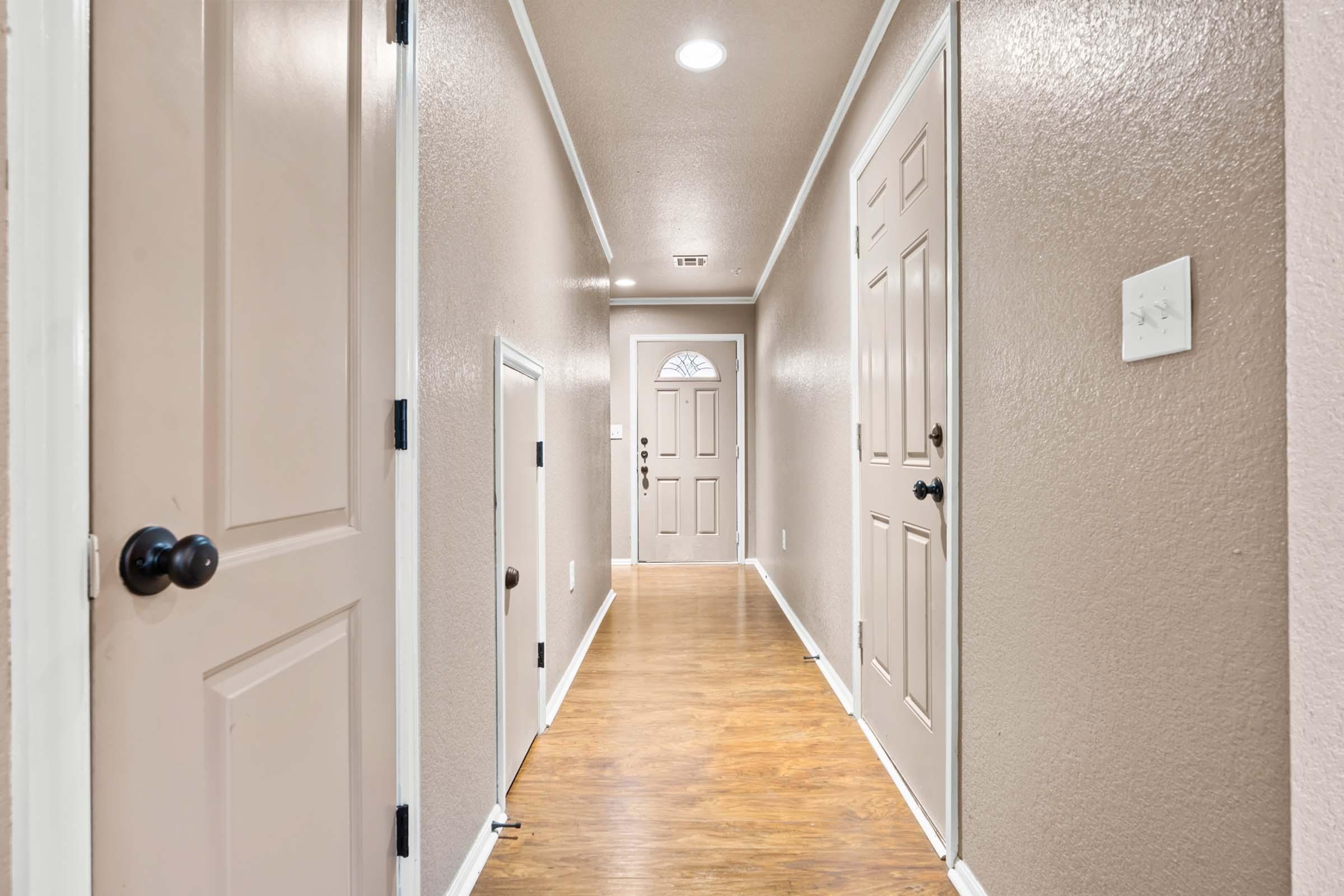 A long hallway with beige walls and hardwood flooring. It features several closed doors on either side, leading to adjacent rooms or closets, and a front door at the far end. Recessed lighting illuminates the space, creating a warm and inviting atmosphere.