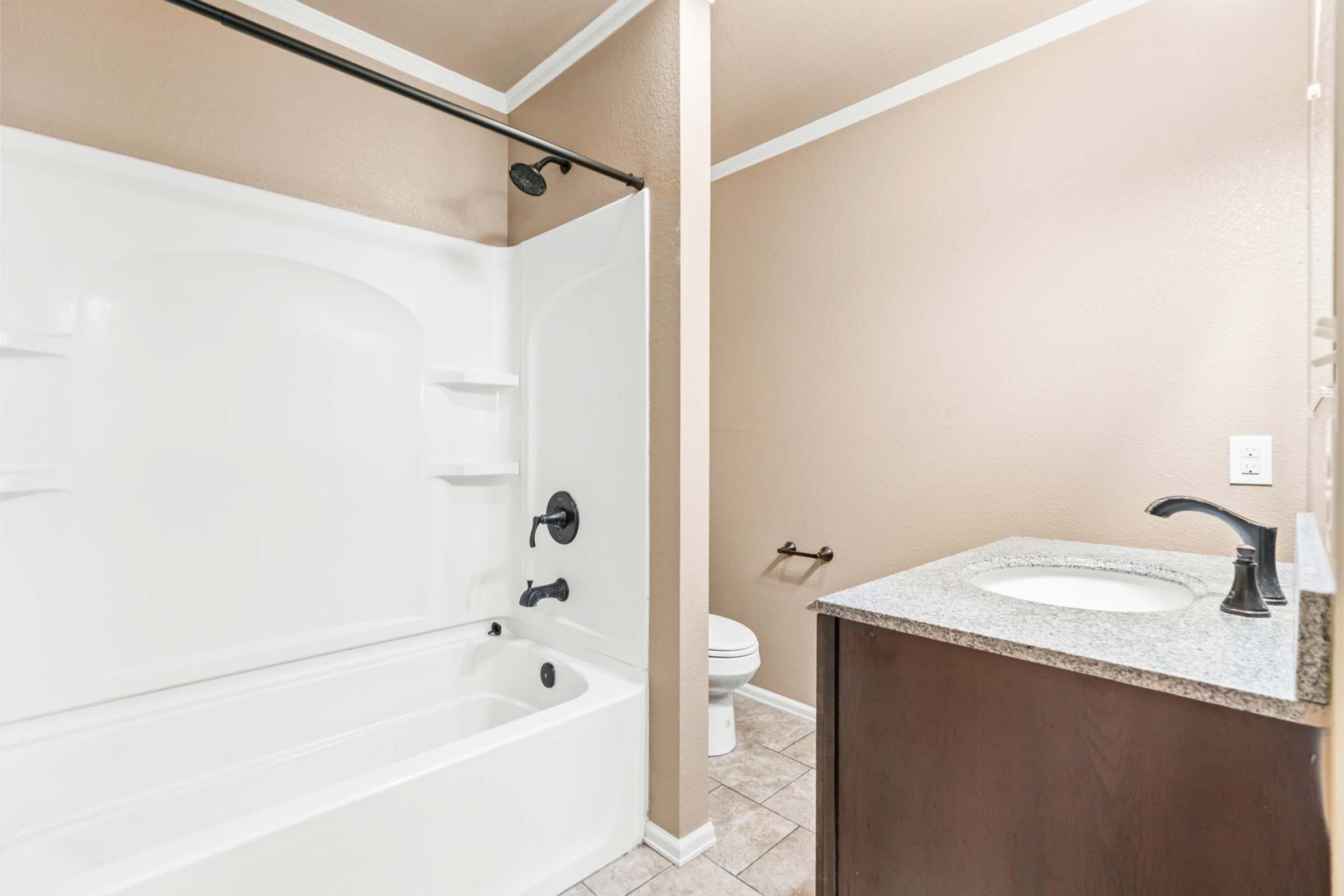 A clean and modern bathroom featuring a white bathtub and shower combination, a dark wood vanity with a granite countertop, a round sink, and a toilet. The walls are painted in a neutral tan color, and there are minimal decorative elements.