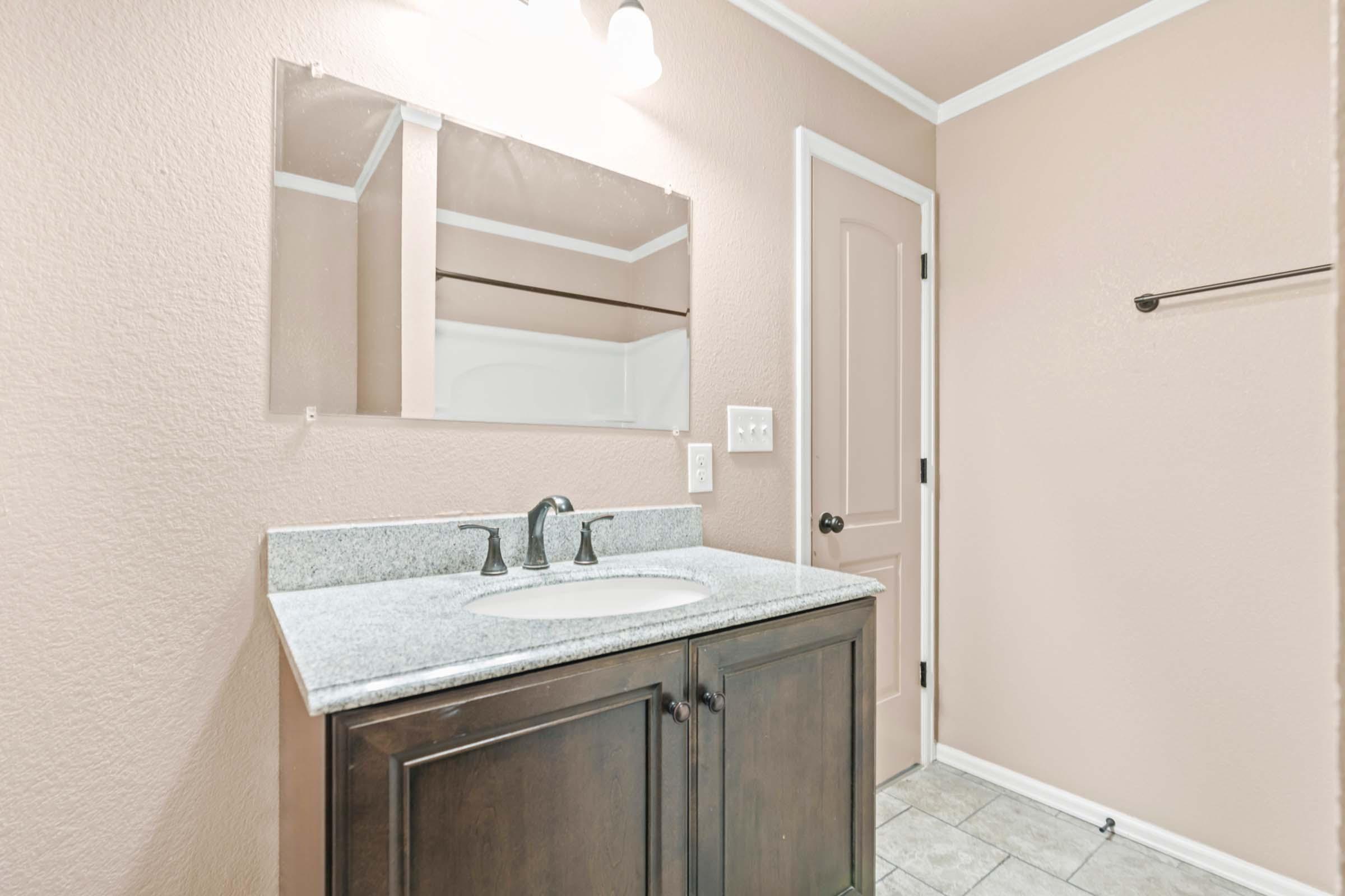 A well-lit bathroom featuring a light pink wall, a dark wood vanity with a white sink, and a large mirror above it. The flooring is tiled, and there's a closed door in a matching pink shade. A towel rack is mounted on the wall beside the door. Overall, the decor is modern and inviting.
