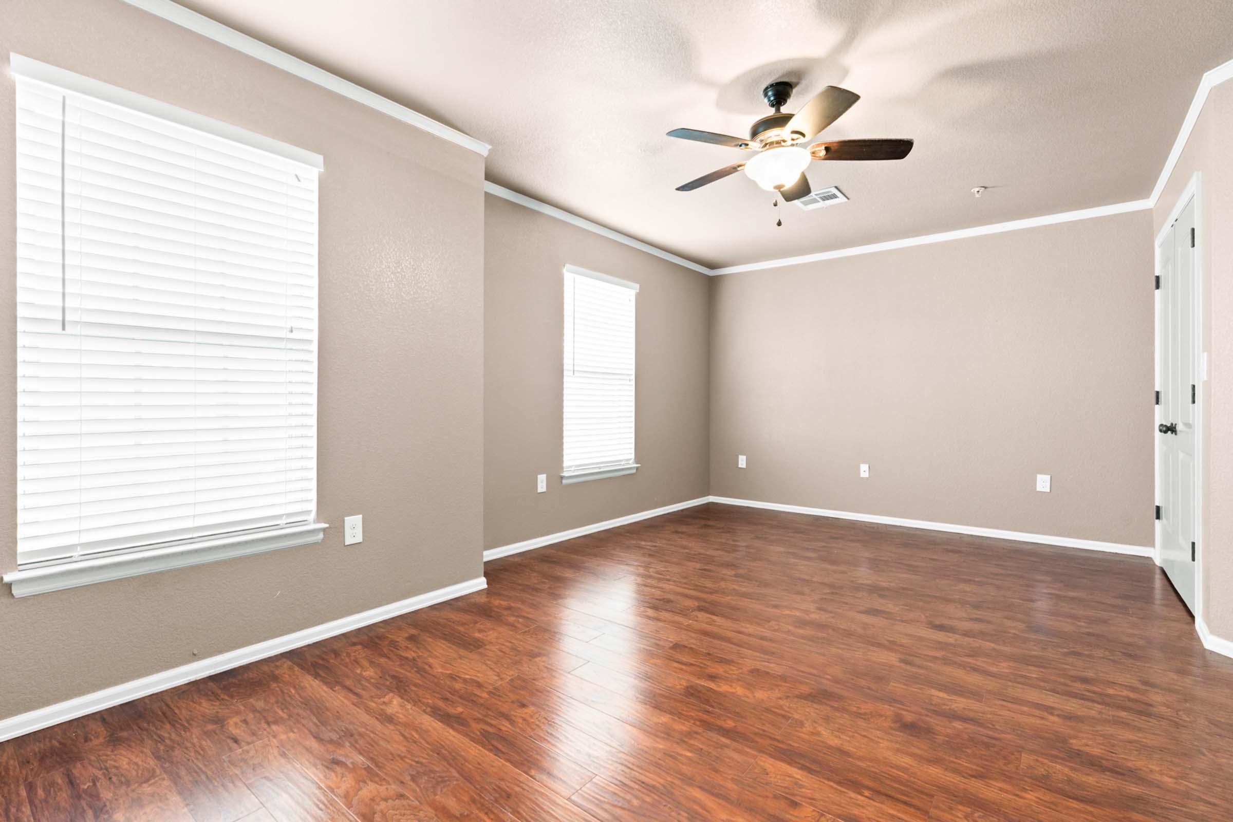 A well-lit empty room with light brown walls and a ceiling fan. The floor is made of wood, and there are two windows with white blinds, allowing natural light to enter. A closed door is visible on the right, adding to the spacious, uncluttered feel of the room.