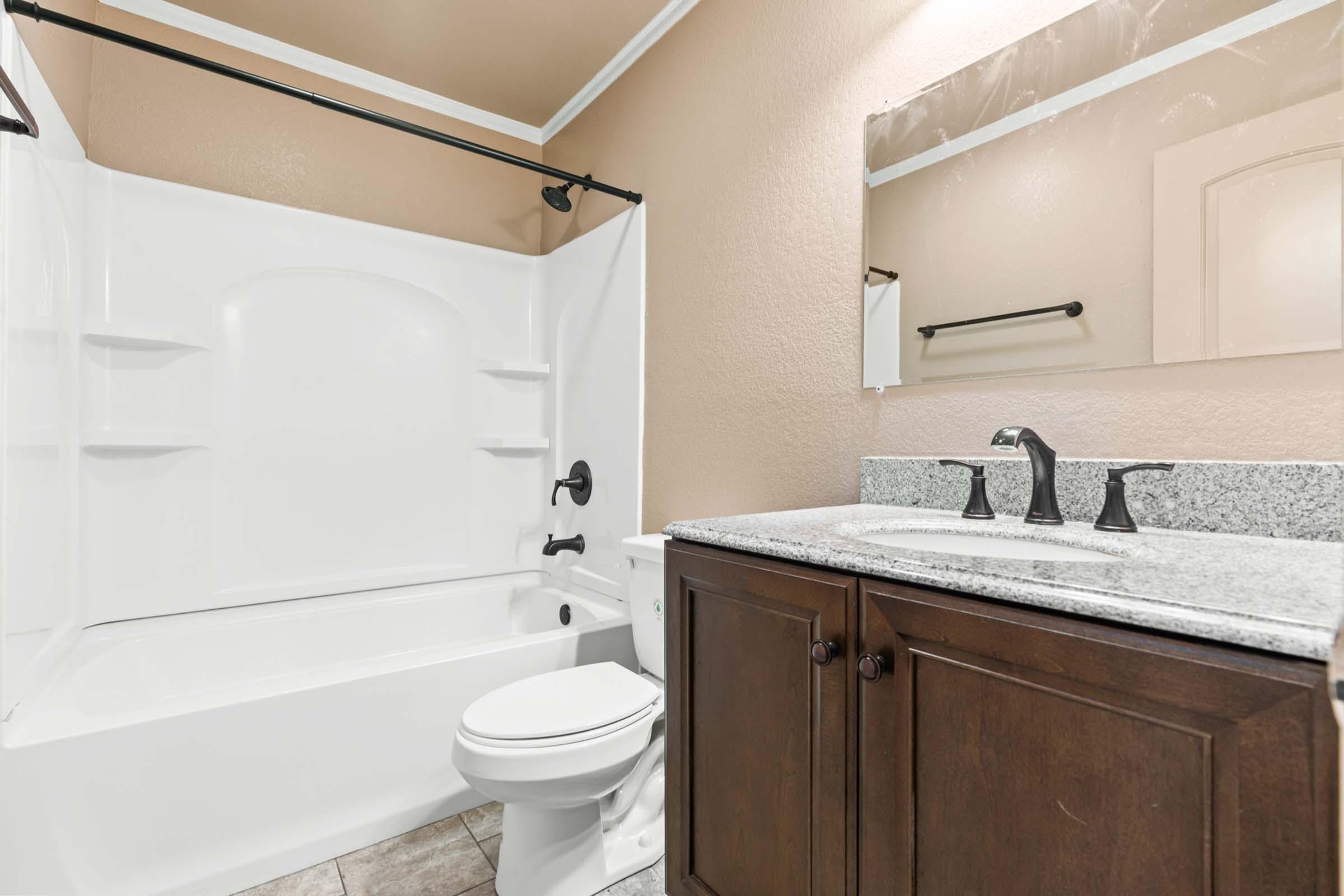 A small bathroom featuring a white bathtub with a shower, a dark wood vanity with a granite countertop, a mirror above the sink, and a neutral-colored wall. There is also a toilet and a towel bar on the wall. The flooring is tiled in a light pattern.