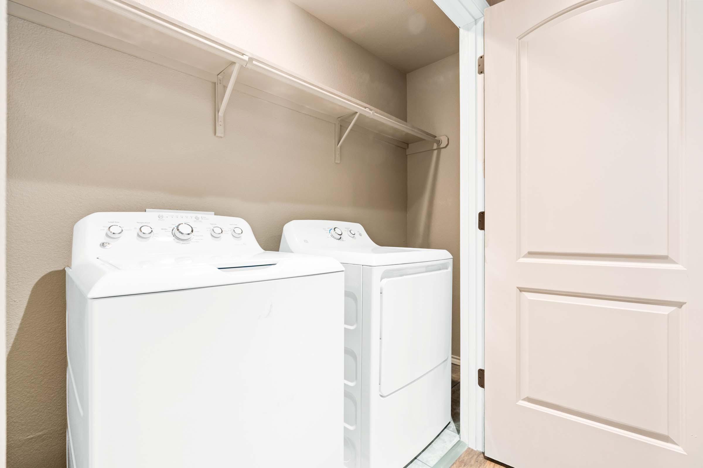 A laundry room featuring a white washing machine and a dryer side by side. The machines are placed against a light-colored wall with a shelf above them. A closed door is visible on the right side, creating a cozy and functional laundry space.