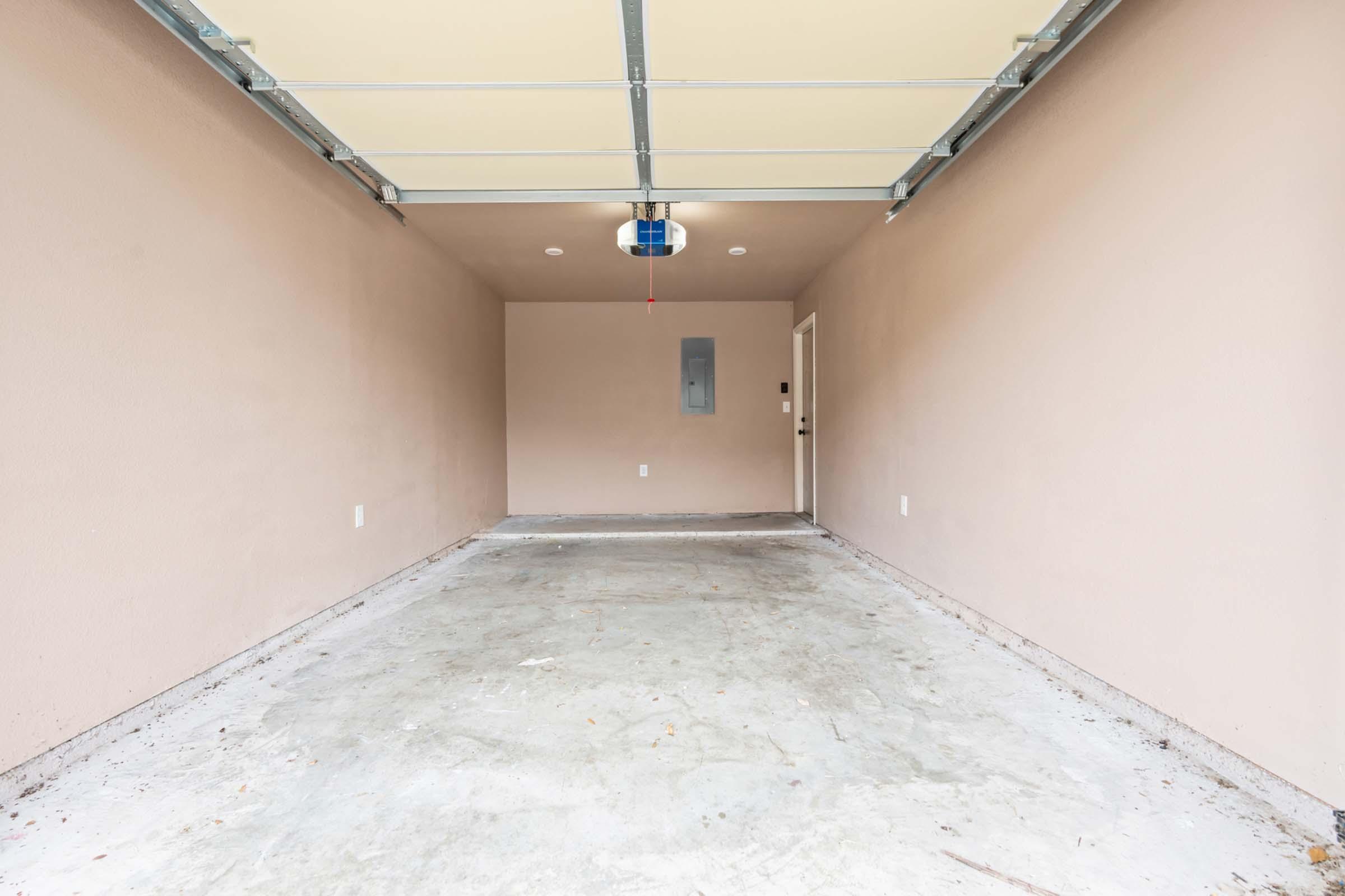 A clean, empty garage with plain beige walls and a concrete floor. The ceiling features a garage door mechanism, and there is a light fixture in the center. Along one wall, there is an electrical panel, and a door leading outside is visible. The space is well-lit and free of clutter.